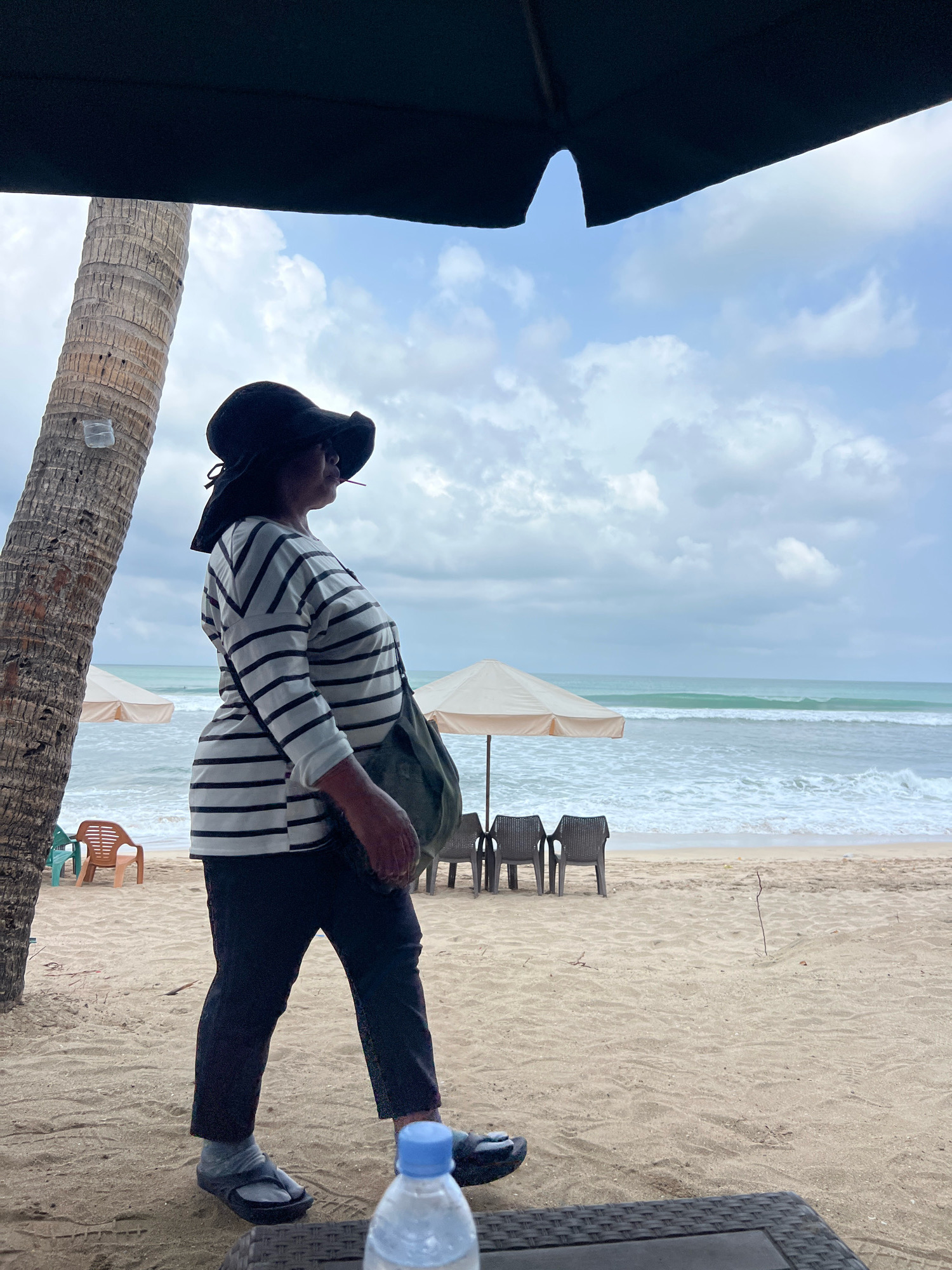 The image depicts a woman walking on a sandy beach in Kuta, Indonesia. She is wearing a black hat, a black and white striped long-sleeved shirt, and dark pants. A green bag is slung across her shoulder. She is looking to her right, away from the camera. In the background, the ocean with gentle waves is visible, along with beach umbrellas and chairs. A palm tree stands to the left of the woman. The sky is cloudy. In the foreground, a table with a bottle of water is partially visible. The overall scene suggests a casual day at the beach.