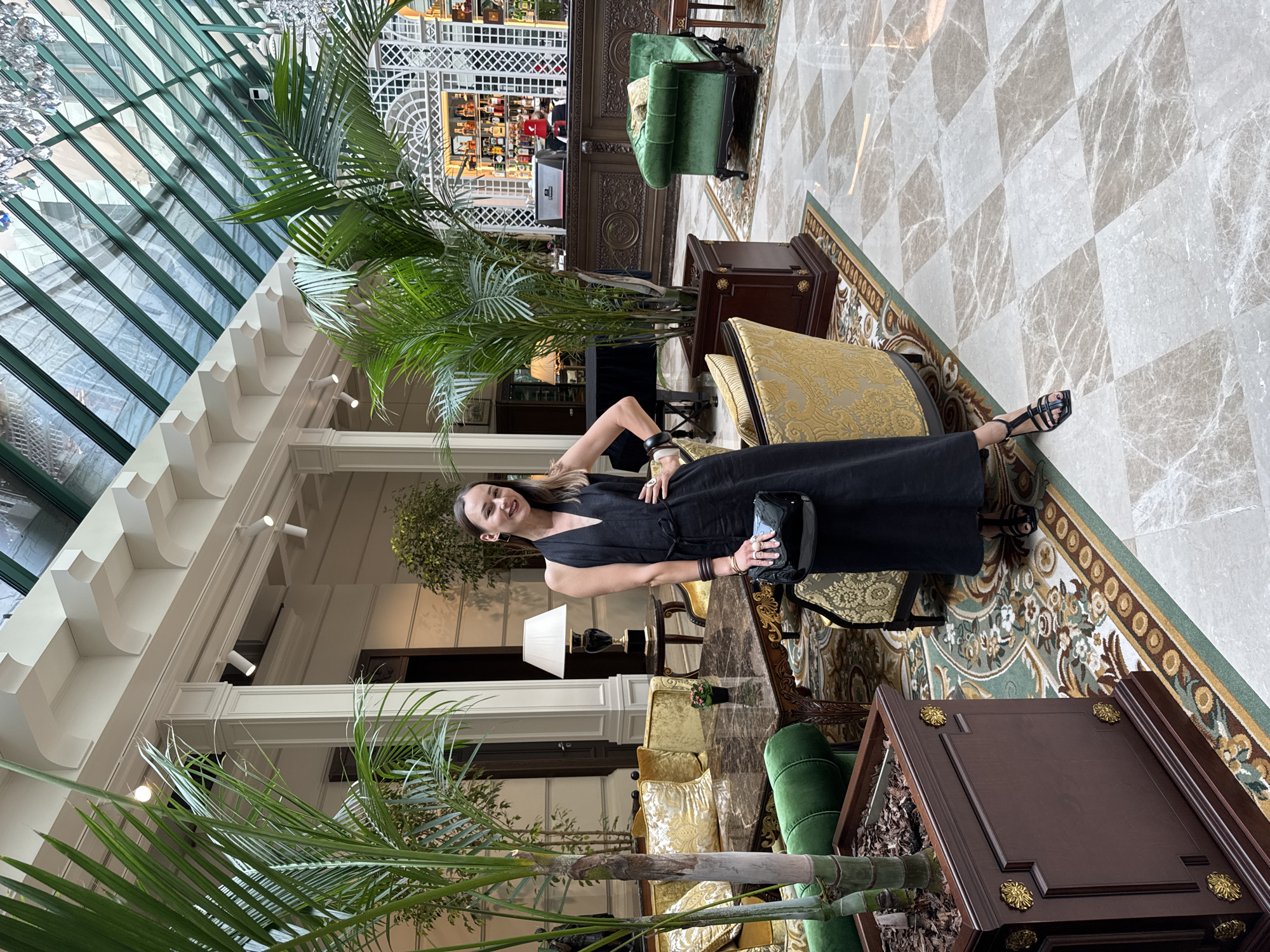 A young woman stands in a grand hotel lobby in Moscow, Russia, wearing a black dress and black sandals. She is holding a black purse. The lobby features ornate furniture, potted plants, and a glass ceiling. A bar area is visible in the background.