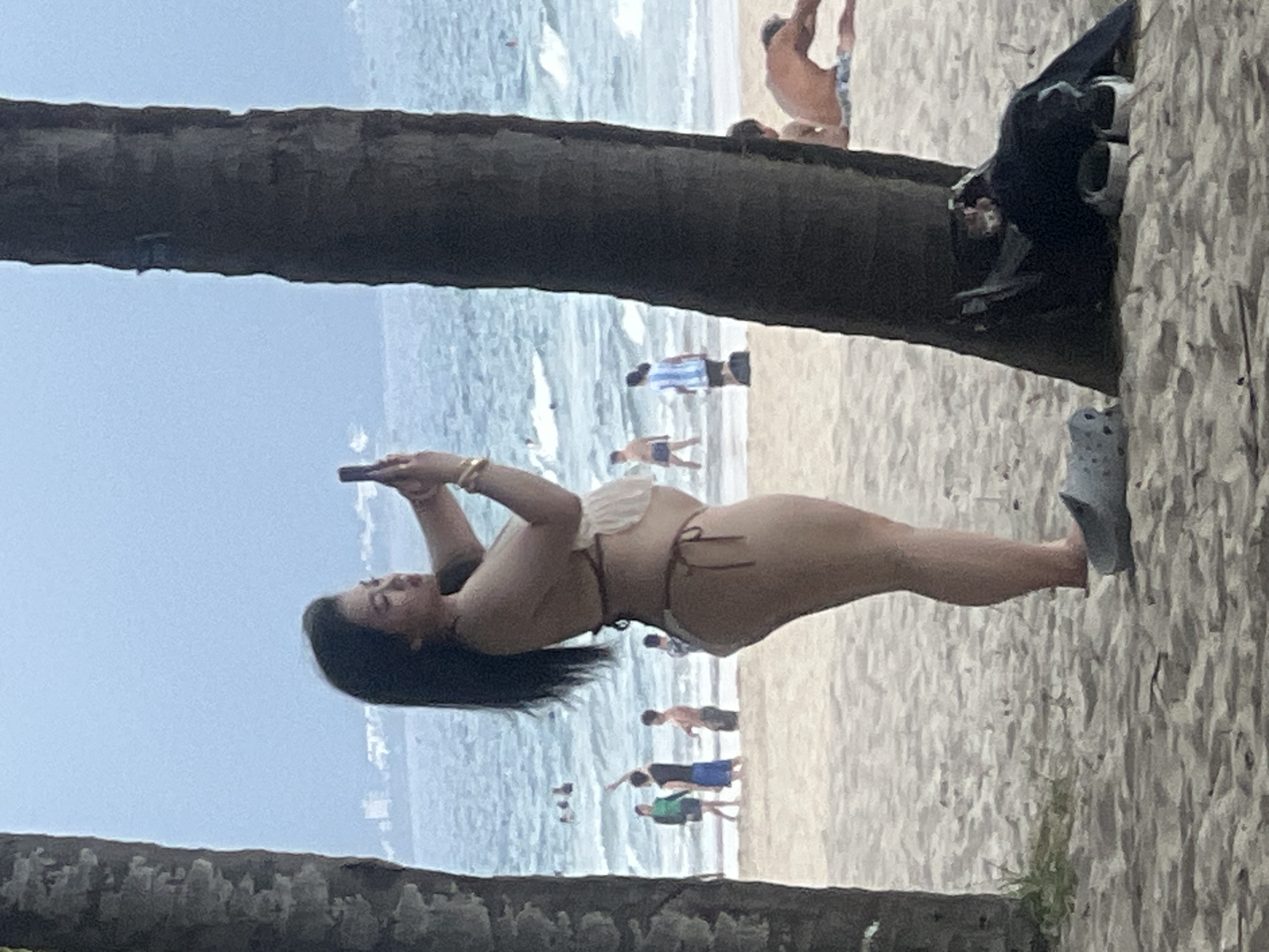 In Da Nang, Vietnam, on a sandy beach, a woman in a bikini stands between two palm trees, taking a photo with her smartphone. She is positioned in the foreground, facing the ocean, with her back slightly turned towards the viewer. The ocean is visible behind her with gentle waves and several people wading or swimming in the distance. The sky is overcast. In the background, a faint outline of buildings suggests a coastal city. In the immediate foreground, near the base of one of the palm trees, a dark bag and a pair of sandals are placed on the sand.