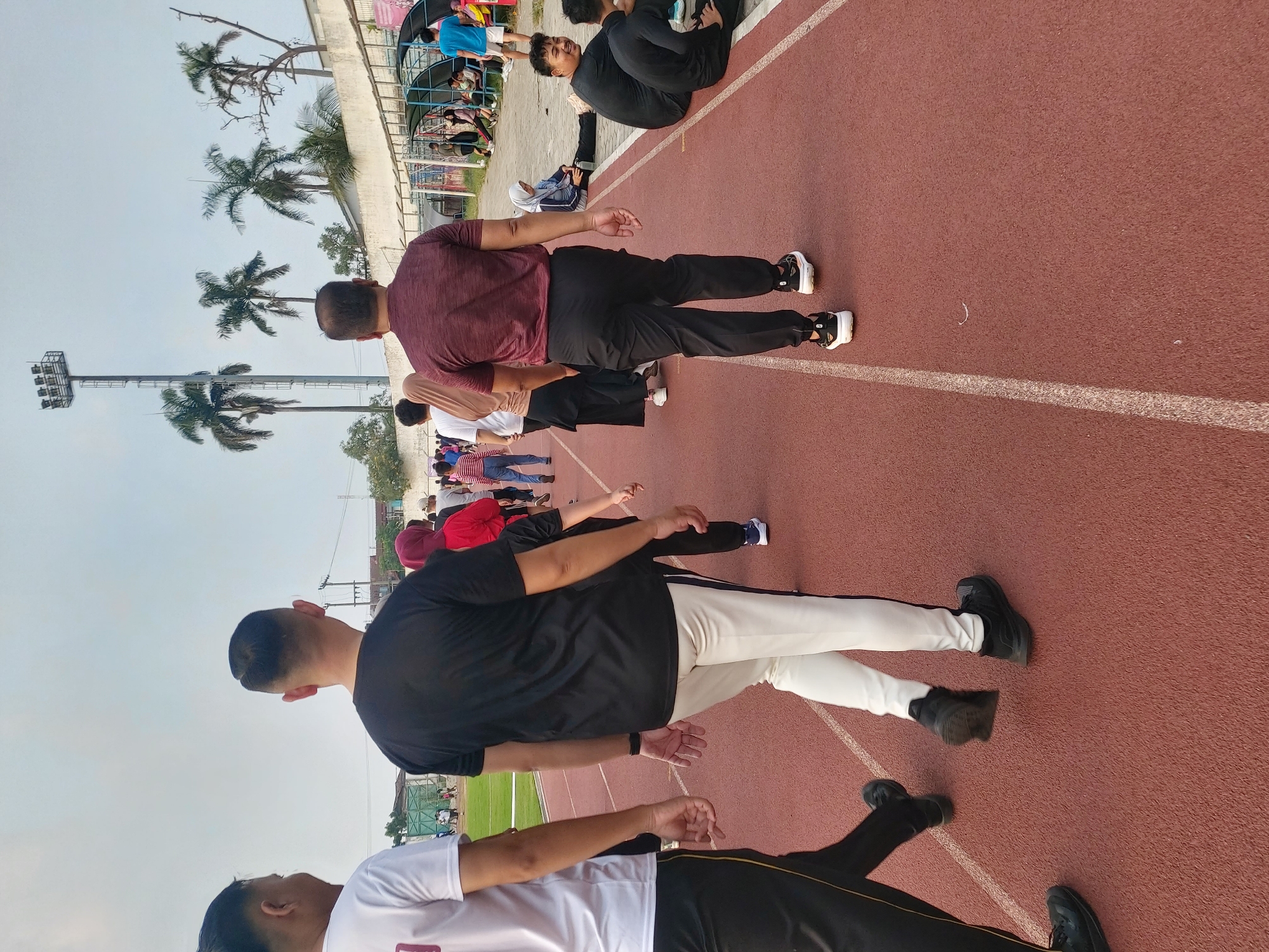 The image shows a group of people walking on a red running track. In the foreground, two men are walking away from the camera. The man on the left is wearing a white t-shirt and black pants with a yellow stripe. The man on the right is wearing a black t-shirt and white pants with a black stripe. Behind them, other people are walking, including a woman in a red headscarf and a woman in a beige headscarf. In the background, there are palm trees, a stadium light tower, and a white building with a blue-roofed structure. Some people are sitting on benches under the blue-roofed structure. The scene appears to be outdoors at a sports facility. This sports facility is located in Bandar Lampung, Indonesia, likely within a larger park or recreational area.