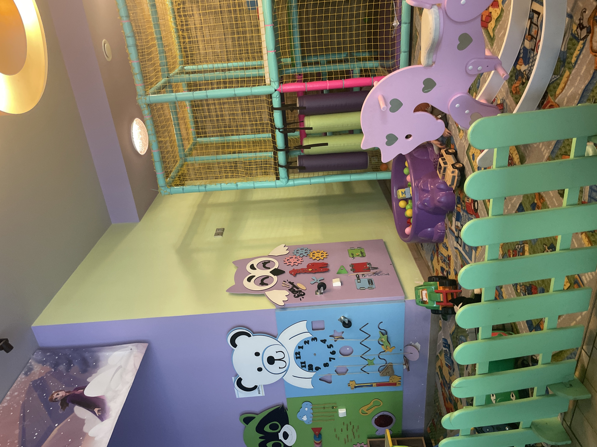 The image shows an indoor children's playroom. The room is painted in pastel colors: purple, green, and light gray. A multicolored play structure with netting is in the background, partially obscuring a wall covered in a carpet with a street-themed pattern. A light purple rocking horse and a purple ball pit are visible to the right of the play structure. The right foreground is dominated by a light green wooden fence.
On the left wall are three play panels featuring different designs: an owl with gears, a bear with a clock, and a raccoon with a slide.
The location is possibly a playroom in Izhevsk, Russia, but this cannot be confirmed definitively from the image alone.