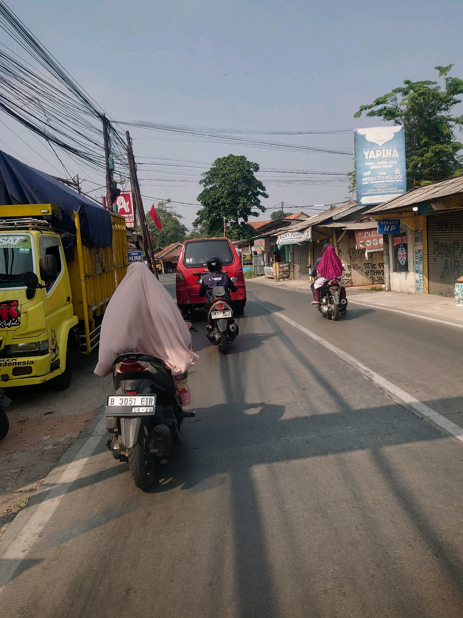 The image depicts a street scene in Bojong Gede, Indonesia, with a focus on a road carrying vehicles and pedestrians. A bright yellow truck, possibly a delivery vehicle, is parked on the left side of the road, partially obscuring a section of a building. In front of the truck, a motorcycle with a rider wearing a light-colored hijab is visible, with its license plate reading "B 3051 EIR 05-28". Further down the road, a red car is in motion, followed by another motorcycle carrying two individuals, one of whom is wearing a dark purple hijab. The road is paved and has a white lane marker. On the right side of the street, several small shops with corrugated metal roofs are lined up. A prominent sign for "YAPINA" and "SMK MEDIATEKNOLOGI" is visible, indicating an educational institution. Overhead, a complex network of electrical wires stretches across the scene, supported by wooden utility poles. The sky is clear and blue, suggesting a sunny day. Shadows cast by the overhead wires and possibly trees create patterns on the road. The overall impression is that of a typical urban street in Indonesia, characterized by the presence of motorbikes, local businesses, and overhead infrastructure.
