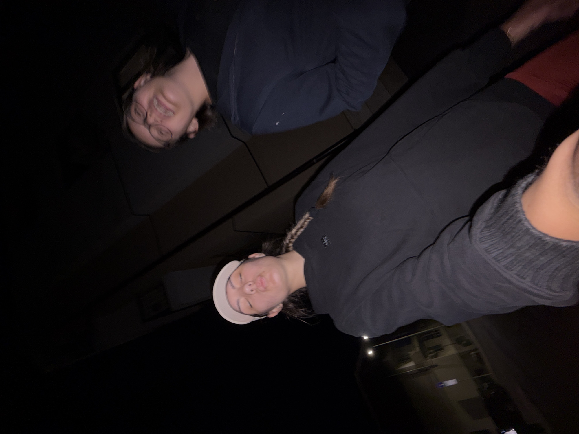 The image shows two young people outdoors at night, with buildings and streetlights visible in the background. The person in the foreground, wearing a black hoodie and a white baseball cap, is making a kissing face with their eyes closed. Their right arm is extended towards the camera, holding it. The second person, wearing glasses and a dark hoodie, is blurred and looking towards the camera with their mouth open. The location is identified as Burgas, Bulgaria. The scene appears to be a street at night.