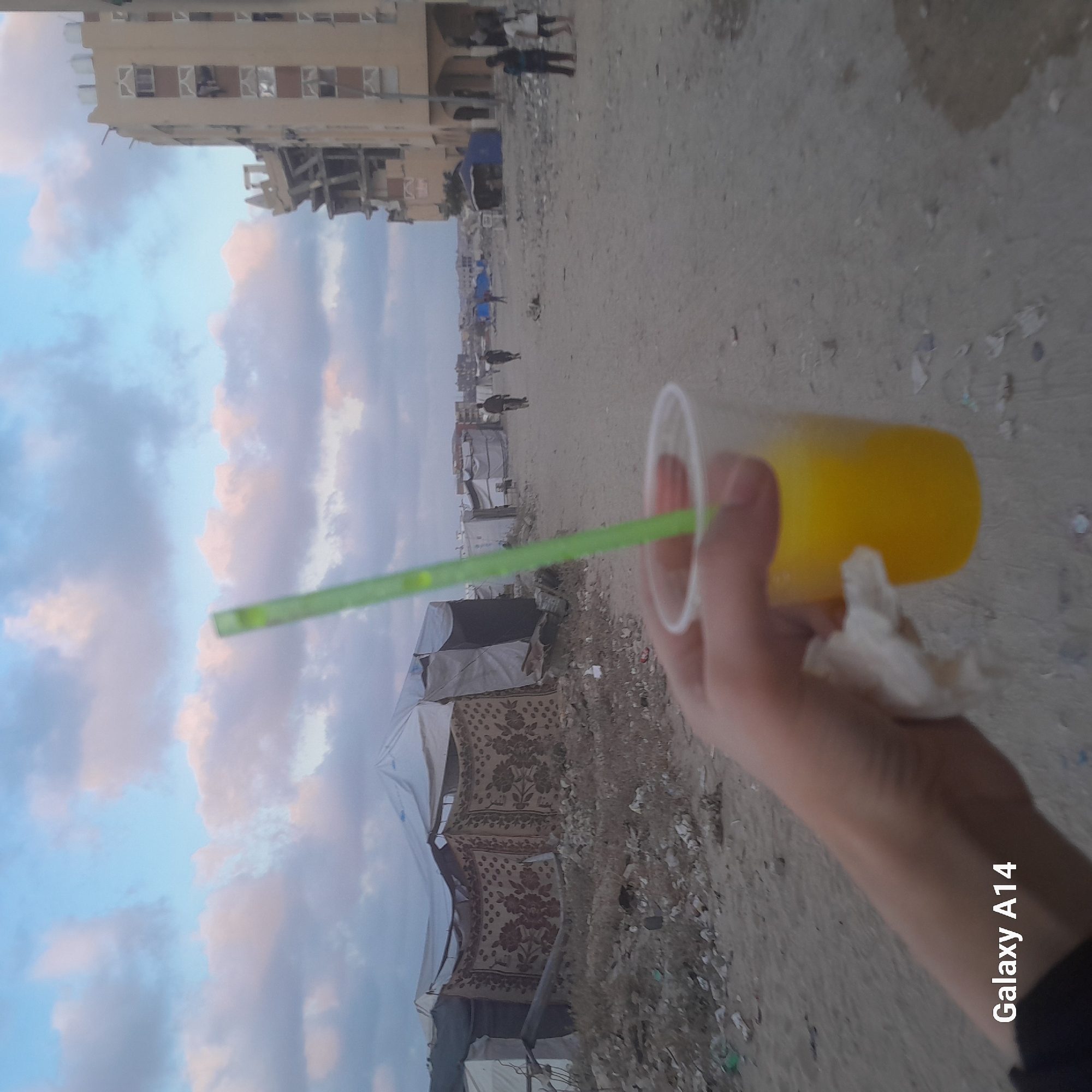 The image shows a person holding a plastic cup with a yellow beverage and a green straw. In the background, there are tents, damaged buildings, and debris, suggesting a war-torn area. The sky is cloudy with hues of blue and pink. There are a few people visible in the distance, some walking and one on a bicycle. The overall scene appears to be in Khan Yunis, Palestinian Territories, an area known for its conflict and displacement. The text "Galaxy A14" is overlaid on the bottom left of the image, indicating it was taken with a Samsung Galaxy A14 phone.