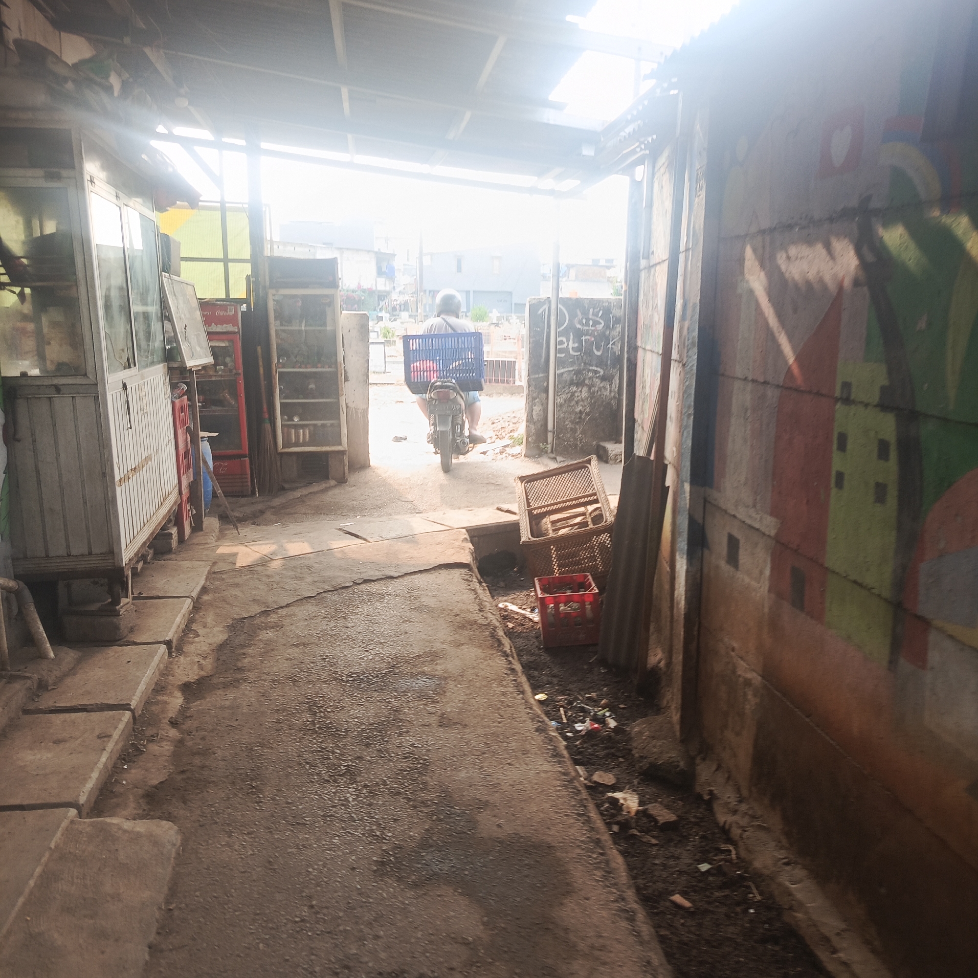 The image depicts a narrow alleyway in a dense urban environment, likely a slum or informal settlement, in South Jakarta, Indonesia. Sunlight streams in from above, creating a bright, hazy atmosphere. On the left, a food stall or kiosk with glass display cases is visible, along with stacked crates, possibly containing beverages. To the right, a rough-textured wall is adorned with vibrant, stylized graffiti depicting abstract shapes and building facades.
In the center of the alley, a person on a motorcycle is seen from behind, wearing a helmet and shorts, and carrying a large blue plastic crate on the back of the bike. The motorcycle appears to be moving away from the viewer, towards the light. The ground is uneven, with a mix of dirt, gravel, and paved sections, and some debris is scattered along the edges of the alley. In the background, residential buildings and structures typical of densely populated urban areas can be seen. The overall impression is one of a bustling, functional, and somewhat chaotic local community.