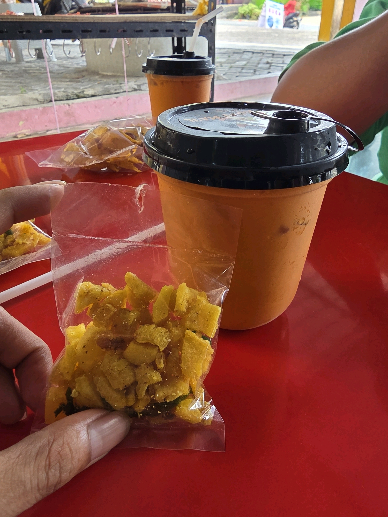 An eye-level, close-up, medium shot captures a hand holding a small plastic bag filled with yellow snacks, which appear to be fried crackers or chips, on a vibrant red table. To the right of the bag, a large orange cup with a black lid and straw is partially visible, suggesting a beverage. The background is blurred but shows a light-colored structure, possibly a stall or food stand, with metal bars and hanging hooks. The environment seems to be outdoors, with hints of natural light and a suggestion of a street or market setting. The context suggests a casual snack and drink purchase at a food stall in Tanjung Pinang, Indonesia.