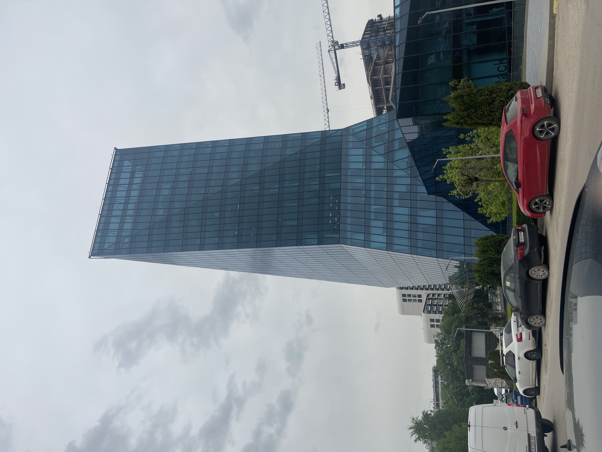 The image shows a street scene in Sofia, Bulgaria. Dominating the view is a modern, glass-facade office building with a unique, angular design. Beside the building, construction appears to be in progress, with a crane visible in the background. A row of parked vehicles lines the right side of the frame. These include a red hatchback, a black sedan, and a white van. Trees and shrubs are visible along the sidewalk, and other buildings can be seen in the distance. The sky is overcast. The image appears to have been taken from inside a vehicle, as part of the dashboard and side mirror are visible in the lower-right corner.
