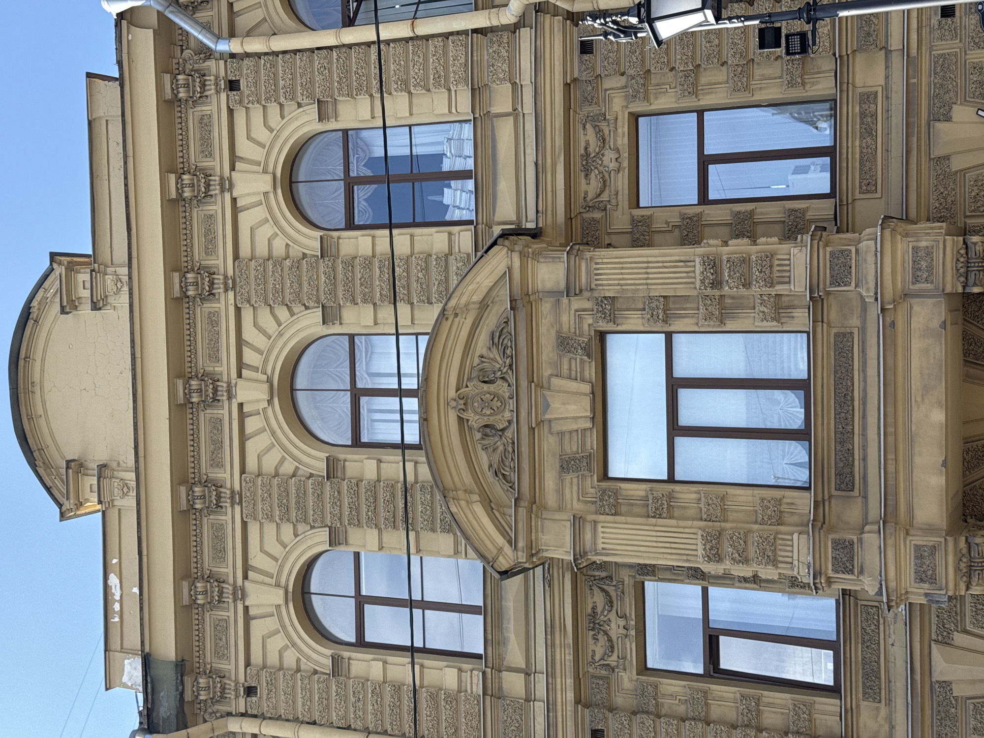 A building facade is shown from a low angle against a clear sky. The facade is made of weathered, yellow stucco with ornate architectural details. The upper floors feature arched windows with brown frames, some with white curtains. Below these, a central, decorative balcony or projecting window area is visible, also with arched openings. The lower floor has rectangular windows, some with reflections of buildings. Architectural ornamentation includes carved friezes, pilasters, and decorative motifs above the windows. A downspout runs vertically along the right side of the building, and a lamppost with attached lights is visible in the foreground.