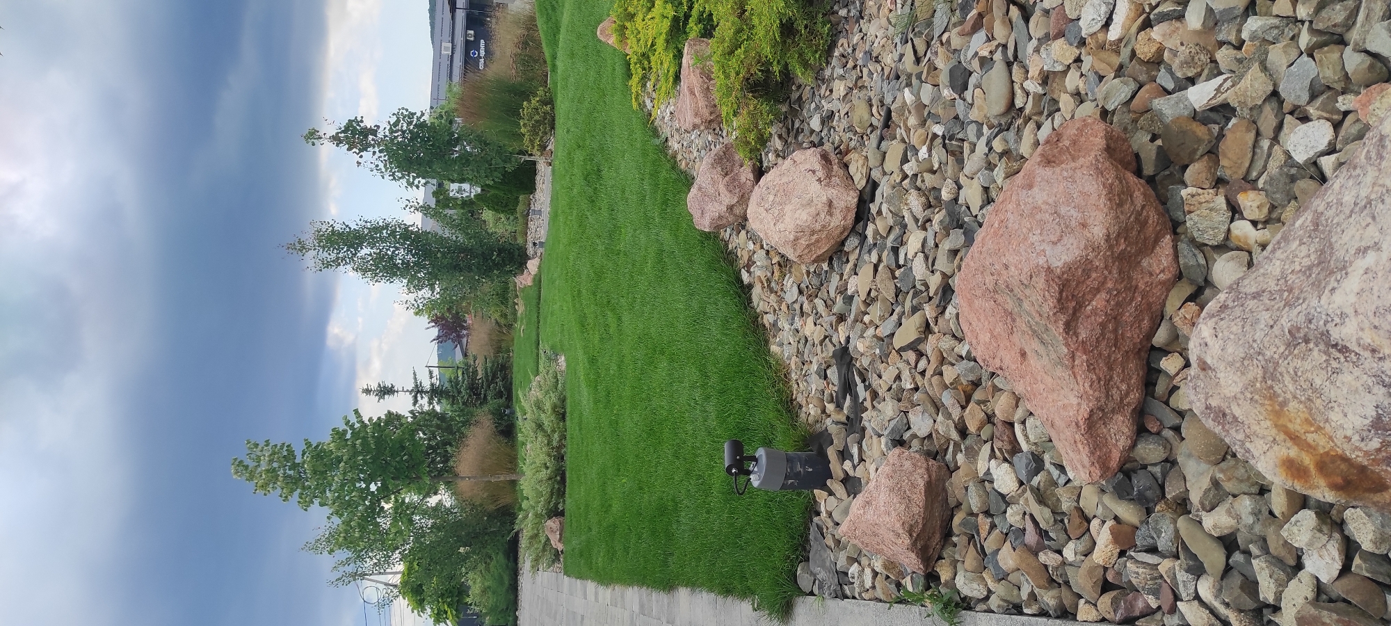 The media file displays a landscape scene at ground level, featuring a neatly mown green lawn bordering a decorative rock garden. The rock garden is composed of various sizes of stones, with several large, reddish-pink boulders interspersed among smaller, multi-colored pebbles. A dark, low-profile garden light is visible partially buried in the gravel, near the edge of the lawn.
Behind the lawn and rock garden, a line of trees and shrubs is visible. Some of the trees are slender with light-colored bark, while others are denser with green foliage. In the background, there is a building with some signage and what appears to be a paved walkway or driveway. The sky is overcast with dark, moody clouds, suggesting a late afternoon or evening on a potentially stormy day. The overall setting appears to be a landscaped outdoor area, possibly a park, garden, or commercial property. There is no clear indication of any people or specific activity taking place. The location context provided is Tulskiy, Russia.