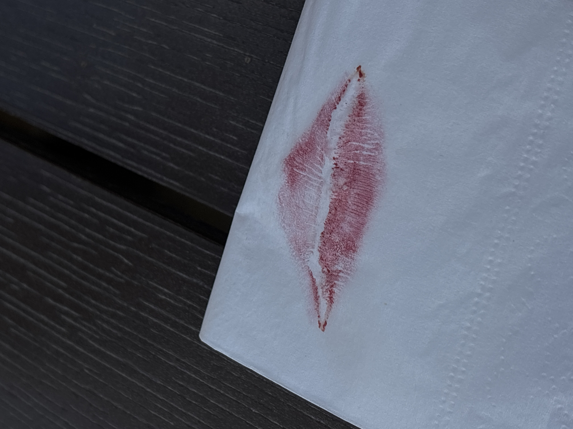 A lipstick print is visible on a white paper napkin, which is resting on a dark brown wooden surface, possibly a table. The lipstick print is a dark red color and diamond shaped. The location is not discernible from the image.