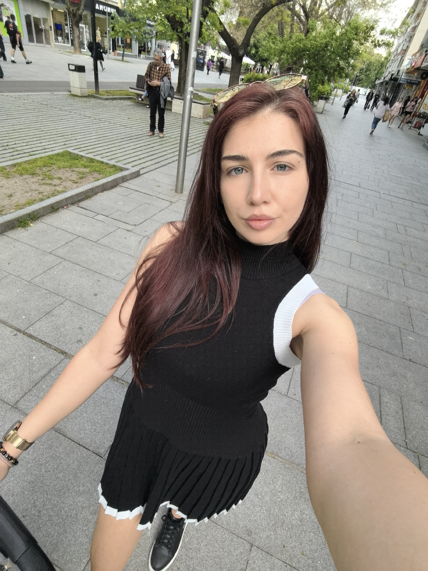 A woman takes a selfie on a city street in Stara Zagora, Bulgaria. She has light skin, blue eyes, and auburn hair. She is wearing a black dress with white trim and black sneakers. She has a gold watch and bracelets on her left wrist. In the background, people are walking on the sidewalk and there are shops and trees. A man in a plaid shirt is walking across the street. The street is paved with gray tiles. Sunglasses rest on the woman's head.