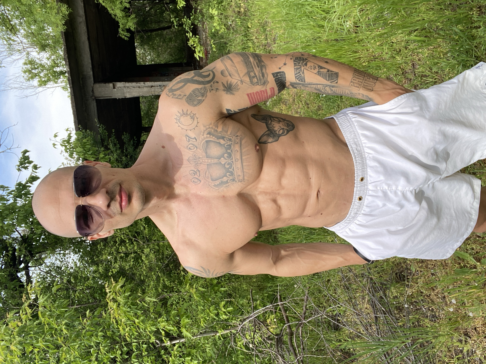 The image is a full-body shot of a muscular, bald man wearing sunglasses and white shorts, positioned in an outdoor, overgrown area. The man has numerous tattoos covering his arms, chest, and abdomen. In the background, there are trees and a dilapidated concrete structure. Although the precise location within Pleven, Bulgaria, cannot be definitively determined from the image, the presence of overgrown vegetation and what appears to be abandoned infrastructure suggests a neglected area.