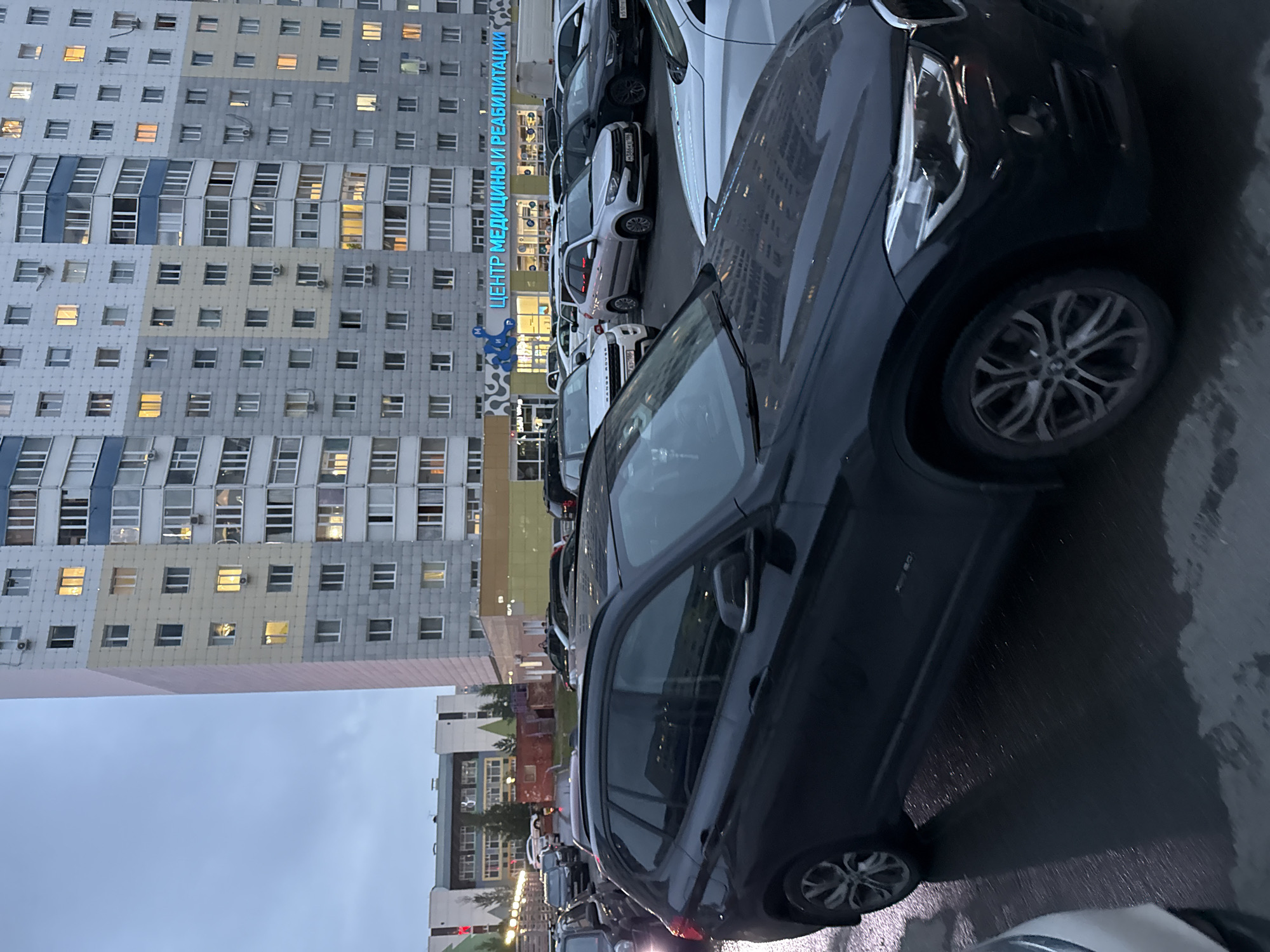 A tall, modern apartment building dominates the background of this outdoor scene, with many windows illuminated from within. In front of the building, a sign in blue neon lettering reads "ЦЕНТР МЕДИЦИНЫ И РЕАБИЛИТАЦИИ," translating to "Center of Medicine and Rehabilitation." A variety of cars are parked in a lot in front of the building. The foreground is occupied by the side and front of a dark-colored SUV, which appears to be in motion or recently parked on a wet surface. The sky is overcast, suggesting it may be dusk or a cloudy day.