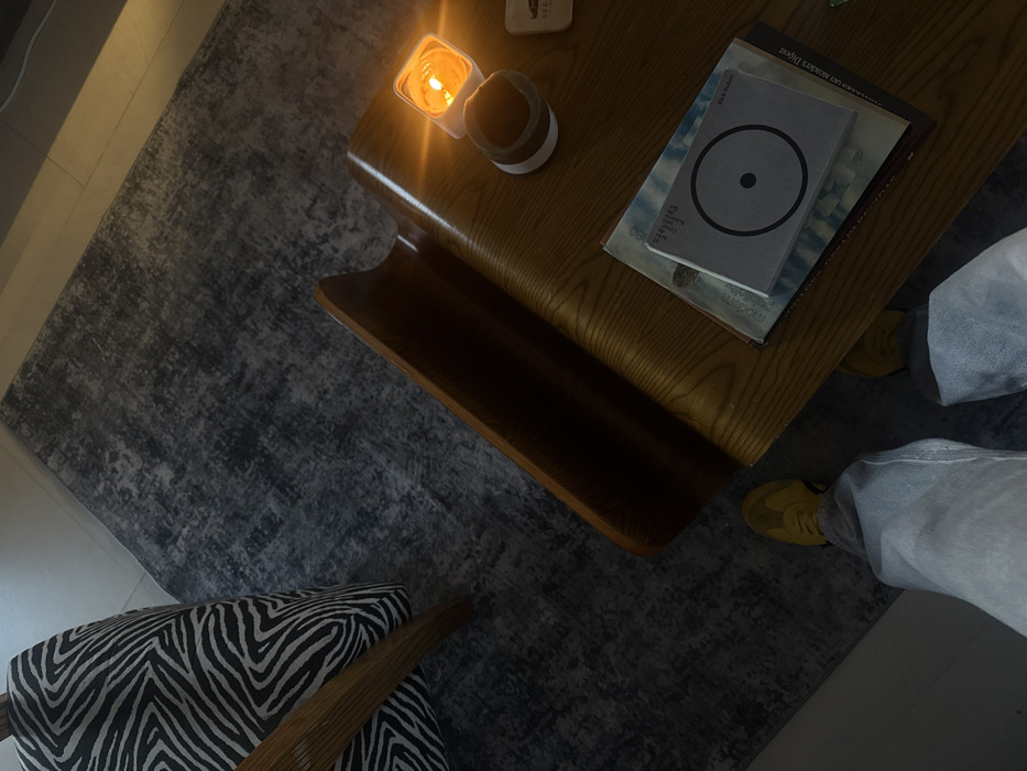 Overhead shot shows a coffee table on a gray rug with a zebra-patterned chair nearby. On the table are a lit candle, a cup, and a stack of books. The topmost book has a gray cover with a black circle on it. A person's legs are visible to the right of the table, wearing yellow sneakers and light-colored pants. The lighting is soft and warm.