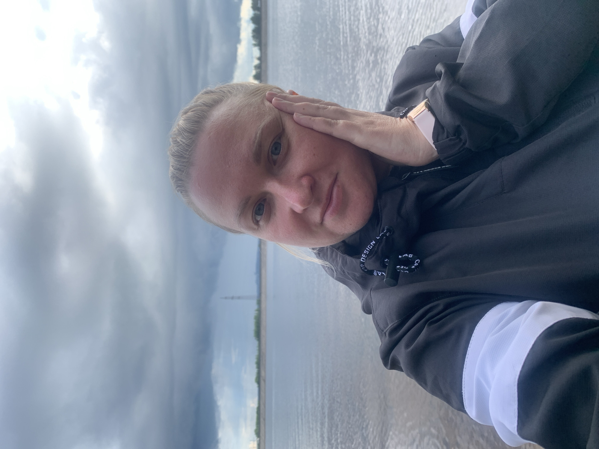 A blonde woman is featured in a close-up, horizontally oriented selfie. Her head is on the left side of the frame, and she gazes directly at the camera with light-colored eyes. Her right hand gently rests on the side of her face. She is wearing a dark grey or black hooded jacket, which features white stripes on the sleeves and a white drawstring with partially visible black text. A rose-gold colored smartwatch with a light-colored band is visible on her left wrist.
Behind the woman, a wide body of water, likely a river or large lake, stretches across the background from the bottom to the top of the image. The water surface is textured with small ripples, indicating movement. A distant, subtle shoreline with a line of trees and a tall, thin structure, possibly a transmission tower, is visible on the far side of the water. The sky, occupying the upper part of the image, is heavily overcast with dense grey clouds, suggesting cloudy weather conditions. A brighter area in the upper left hints at the sun being obscured by clouds.
The lighting is soft and diffused, typical of an overcast day. The woman's expression is calm and neutral, with a slight hint of a smile. Visible text on her jacket's drawstring includes "DESIGN LAB" and "COD" (possibly part of "CODE"). The letters "Chr" are also partially visible on her smartwatch band. The scene depicts a serene outdoor moment by a large body of water, consistent with locations such as Noviy, Russia.