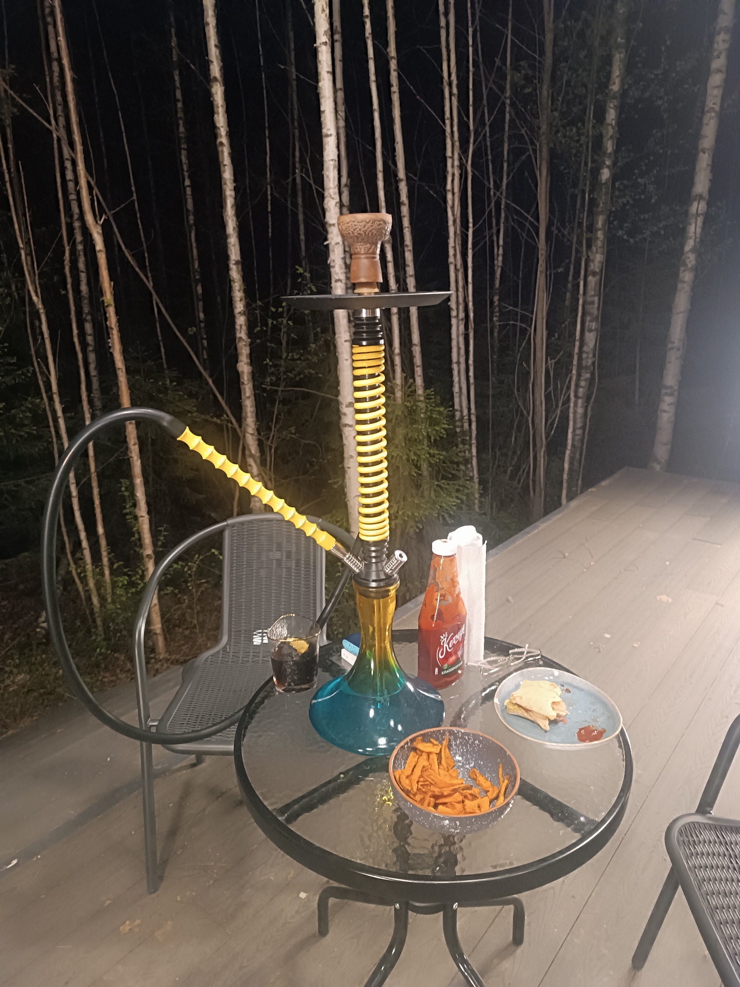 The image shows a hookah with a yellow hose and spiral stem sitting on a black metal patio table. On the table are a bowl of orange fries, a plate with a sandwich and ketchup, a bottle of ketchup, a glass with a dark liquid, and some crumpled napkins. The table sits on a wooden deck. A metal patio chair is behind the table, and there is another chair partially visible on the right. The background features a dark wooded area with tall, slender trees. The scene is lit by an unknown source, giving the impression of night. The location is Unknown, Unknown. There are no people present in the image.