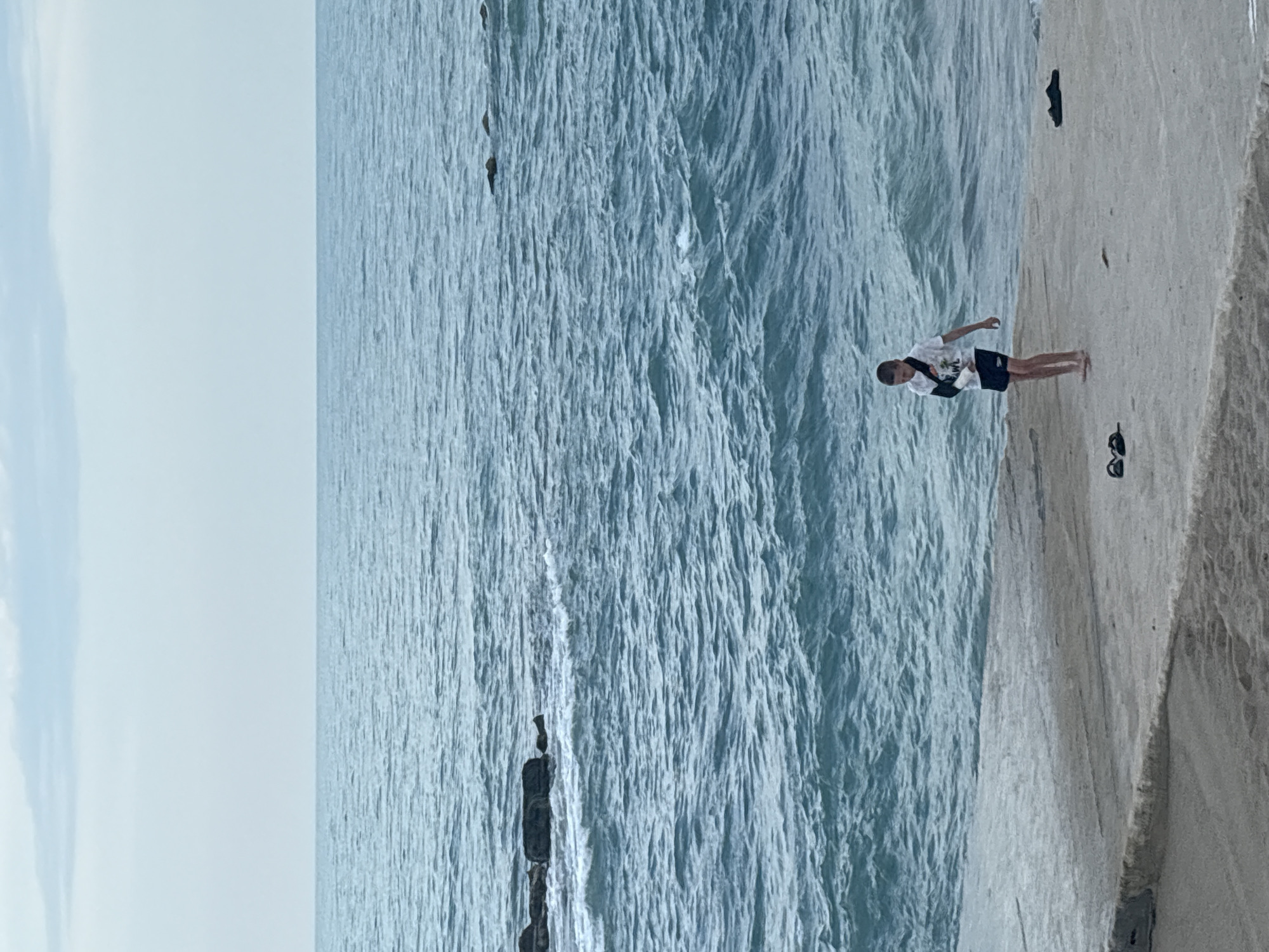 The image, originally rotated, depicts a bright daytime coastal scene. A young person, likely a male pre-teen or teenager, stands on a light-colored concrete or rocky platform that forms the right side of the frame. He is wearing a white t-shirt, dark shorts, and has a black arm sling on his left arm, indicating an injury. His bare feet are visible. Near his feet, on the platform, are a pair of dark sunglasses and another dark object, possibly a shoe.
To the left of the platform, a vast expanse of blue-green ocean or sea stretches towards the horizon. The water is textured with numerous waves and ripples, suggesting movement. Several dark rocks are visible within the water, particularly in the mid-ground. The sky, occupying the top portion of the frame, is a pale, hazy blue with some faint, diffused cloud formations.
The individual appears to be casually standing and observing the water or the distant horizon. There is no specific activity or event taking place beyond observation. The weather seems clear and bright. On the front of the person's white t-shirt, there is faint, dark text or a graphic, but it is not clearly legible from the provided resolution. The overall impression is one of a calm, outdoor moment by the sea.