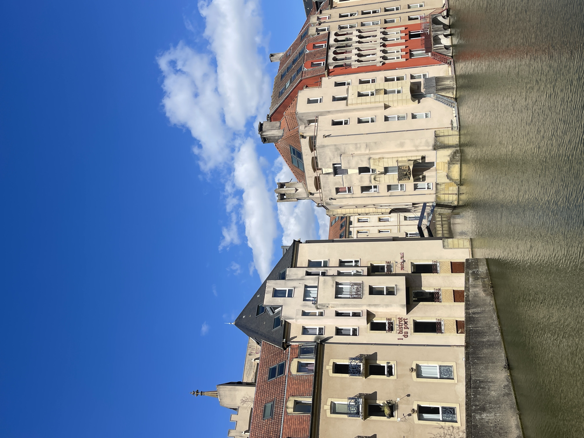 The image presents a view of Metz, France, showcasing buildings along a waterway. The buildings are predominantly light-colored with red-tiled roofs, featuring numerous windows. Some buildings have signage, including "1 bistrot du port" and "restaurant." The water reflects the architecture. The sky is blue with scattered clouds.