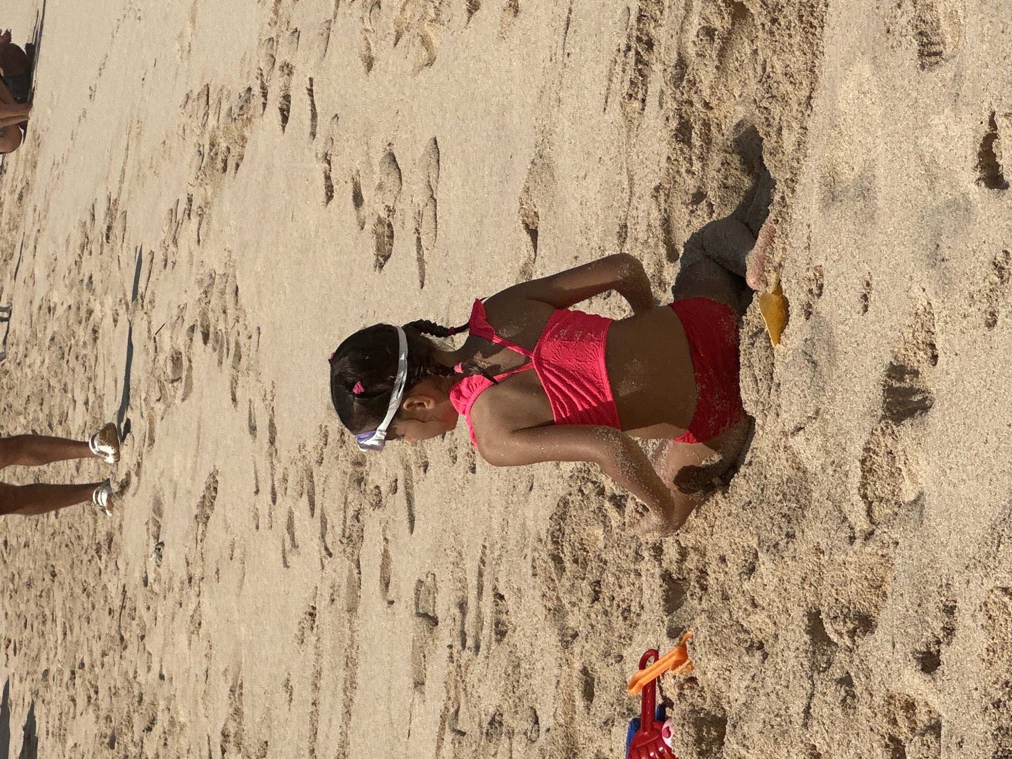 A young girl in a pink swimsuit and goggles is lying on a sandy beach, partially buried in the sand. She has her hair in braids and is looking down. There are footprints in the sand around her. A child's red and orange toy is visible in the lower right corner of the image. The sun is shining, indicating it is daytime. The scene appears to be a recreational activity at a beach in Kuta Selatan, Indonesia, likely during pleasant weather.