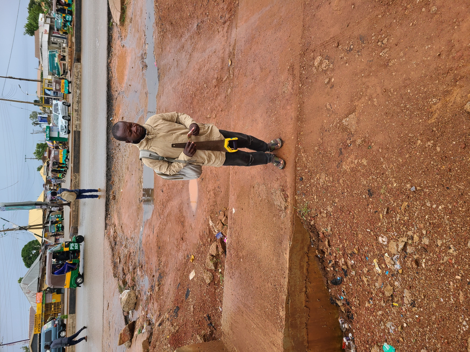 The image captures a man standing on a dirt path in a city, likely Gura Top, Nigeria, holding a saw with a yellow handle and a red object in his other hand. He is wearing a tan jacket, black ripped jeans, and sandals. A backpack is slung over his shoulders. The ground is uneven and covered in red dirt, rocks, and puddles of water, suggesting recent rain or poor drainage. In the background, a street scene unfolds with several auto-rickshaws, a white car, a green truck, and various buildings. The overall environment appears to be a somewhat neglected urban area.