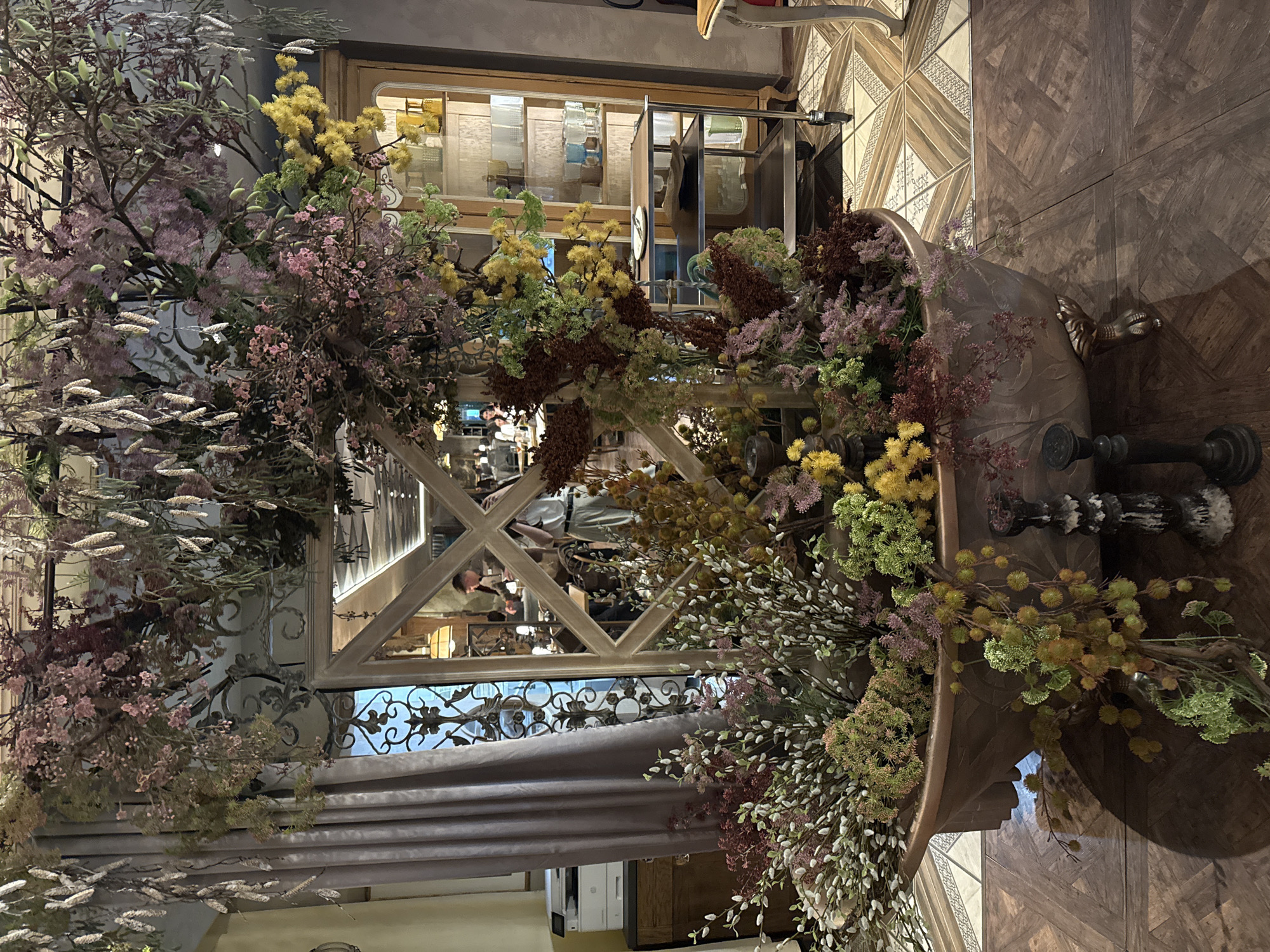 The image shows an indoor scene, likely a decorative floral arrangement in a store or restaurant. A large, ornate vase-like container filled with a variety of flowers and foliage dominates the composition. The floral arrangement features a mix of colors including yellows, greens, pinks, and purples. The background contains a structure with cross-beams, possibly part of the building's architecture or a display fixture. This structure has an ornate iron decoration.
Through the beams of the structure, other interior elements are visible, including what appears to be shelves and a glimpse of people. The flooring has a patterned wooden design. A portion of a gray curtain or fabric is also visible near the bottom left corner. There is no indication about Sofia, Bulgaria in the image.