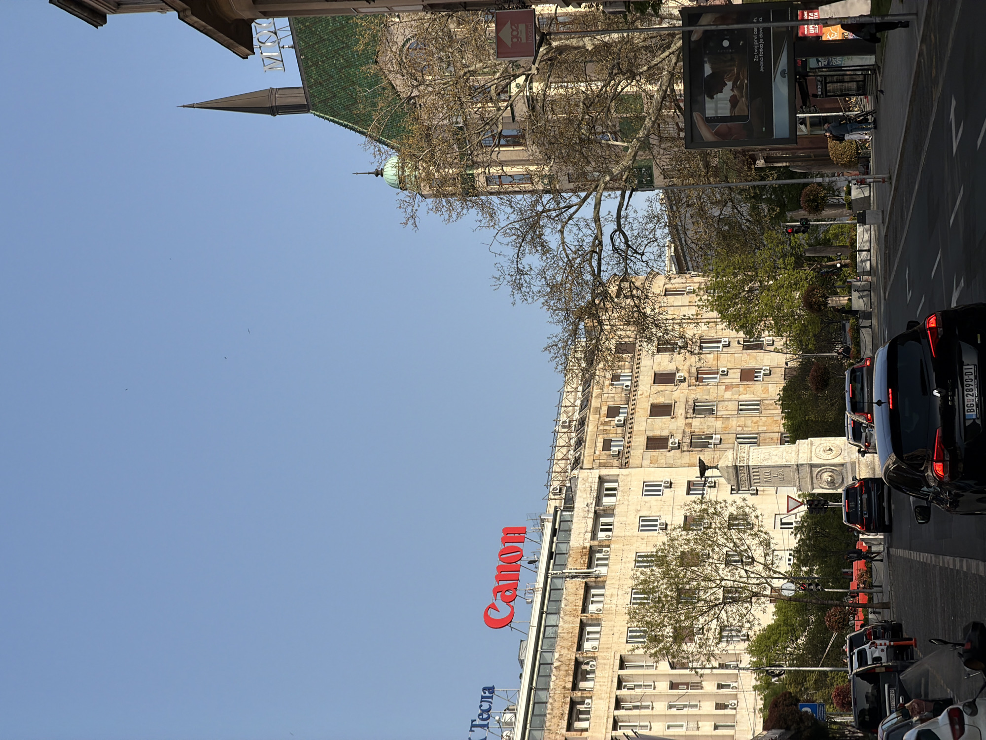 The image shows a street scene in Belgrade, Serbia, dominated by tall buildings. A prominent building features a large "Canon" sign in red letters and a smaller "Тесла" sign in blue. Another building has a green roof and a spire. Traffic is visible, with several cars moving along the street. A billboard stands by the side of the road displaying an advertisement. The sky is clear and blue. A stone monument or structure stands among the buildings, partly obscured by trees.