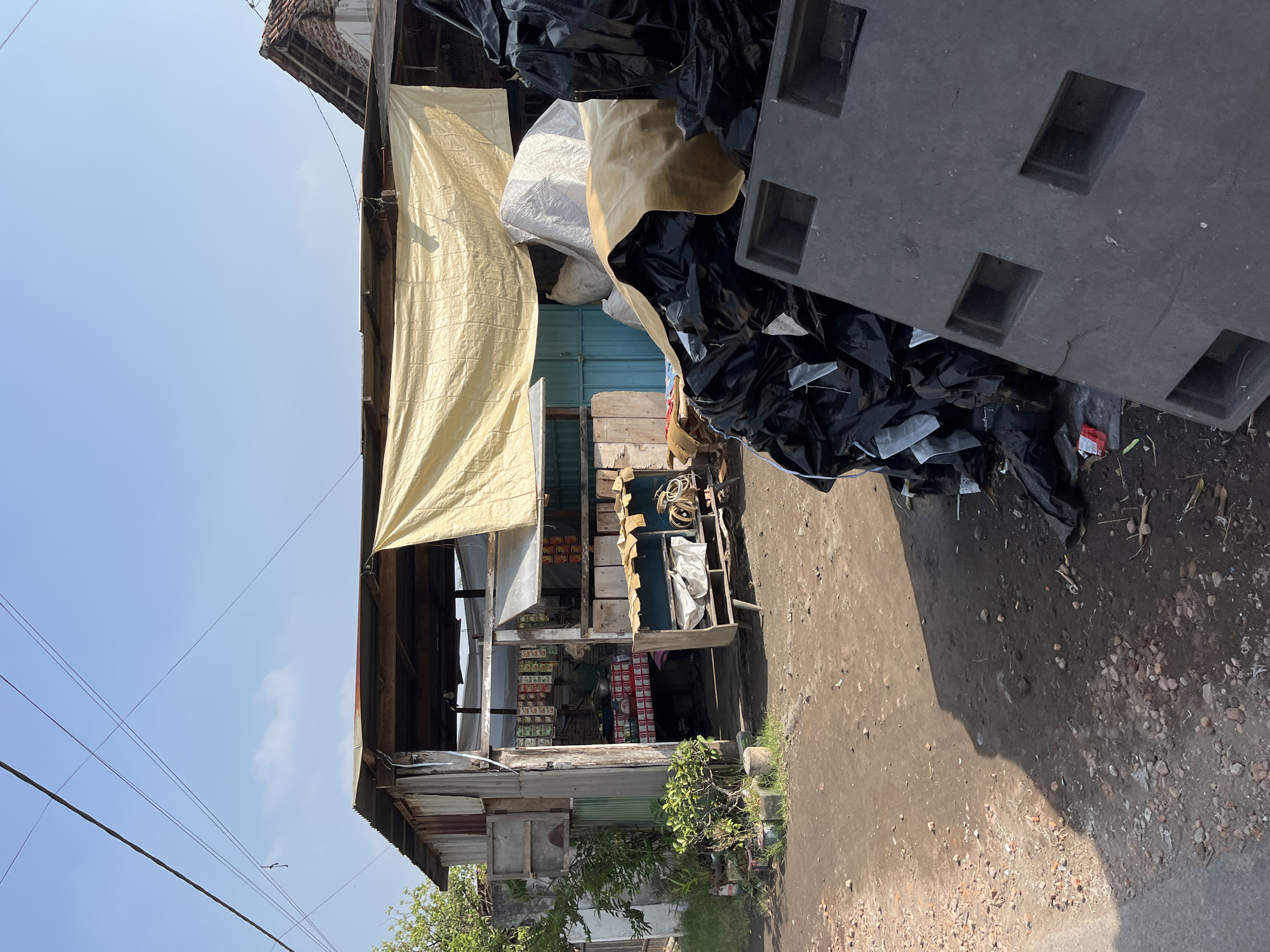The image depicts a street scene in Gempolsari, Indonesia, featuring a small, open-air shop or warung. The structure is made of corrugated metal and wood, with a faded yellow tarp providing shade over the entrance. Inside, shelves are stocked with various packaged goods, and a person, partially obscured, appears to be tending to the shop. In front of the shop, a rustic wooden display stand holds items, including coils of rope and some white bags. To the right of the shop, a large pile of black plastic bags, some appearing to contain trash or recyclables, is stacked near a dark grey, rectangular object with recessed compartments, possibly a pallet or container. The ground is unpaved, with dirt, gravel, and scattered debris. The sky is clear blue with a few wispy clouds, and electrical wires are visible overhead. The scene is characterized by its simple, utilitarian construction and the presence of goods for sale and collected materials, suggesting a typical commercial area in a rural or semi-urban Indonesian setting.