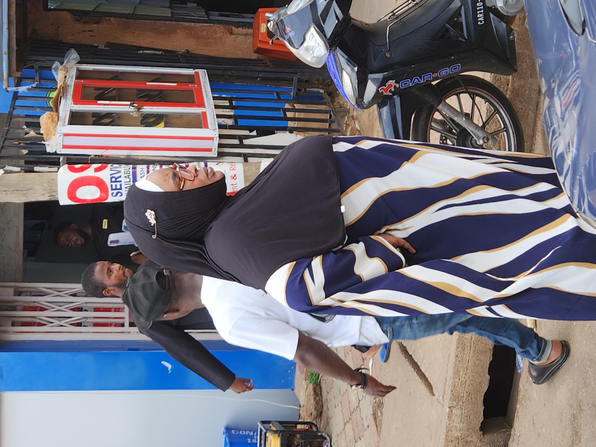 A woman wearing a head covering and a striped dress is standing next to a man wearing a white shirt, jeans, and a black baseball cap. There is a motorcycle with "CAR-GO" on the side, and a blue car or truck next to it. They are on a city sidewalk with a building in the background. The location is likely Unknown, Unknown, based on the presence of the "CAR-GO" motorcycle, which is a common form of transportation in some African cities. There are at least two other men present in the background. One man is looking on at the interaction while the other is wearing a black uniform with his back facing the scene.