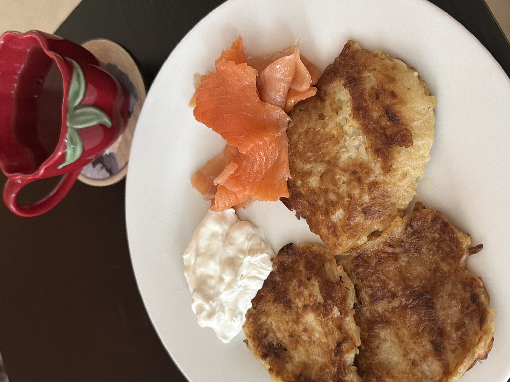 A white plate rests on a dark surface, possibly a table. The plate holds three potato pancakes, a dollop of white sour cream, and several slices of smoked salmon. To the left, slightly out of focus, sits a red, strawberry-shaped mug on a coaster. This image could depict a brunch or breakfast meal in Tel-Aviv, Israel, though the specific location cannot be determined from the image alone. There are no people visible. The focus is on the food and mug, suggesting a culinary theme.