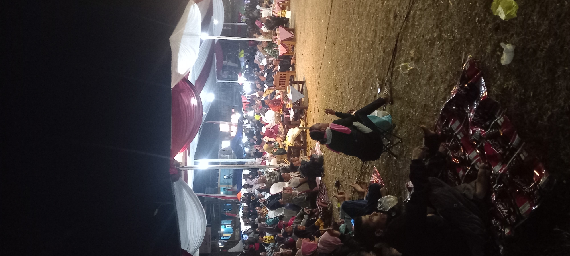 This image depicts an outdoor evening gathering in the city of Wanareja, Indonesia, likely a community event or celebration. The scene is illuminated by artificial lights and a large tent structure covered with red and white fabric, suggesting it might be a formal occasion. A significant crowd of people is seated on the ground, some on mats and others on chairs, facing a central area where a few individuals are seated on low tables and chairs. The audience appears to be watching a performance or listening to a speaker. The ground is covered with grass and scattered debris. The overall atmosphere suggests a lively and engaged community event.