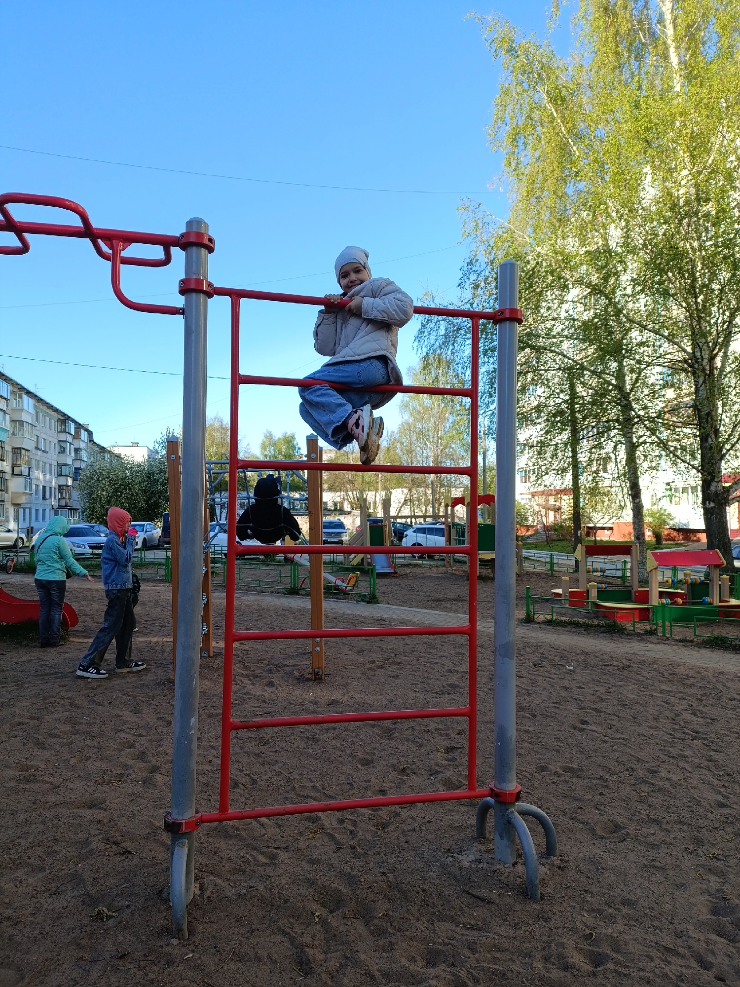 The image shows a playground scene in Chaikovsky, Russia. A young girl in a light jacket, blue jeans, and a beanie sits on the top rung of a red and gray metal ladder structure. Other people, including children and adults, are visible in the background, some near a slide and other play structures. The ground is covered in sand. In the background, there are apartment buildings and trees. The sky is blue.