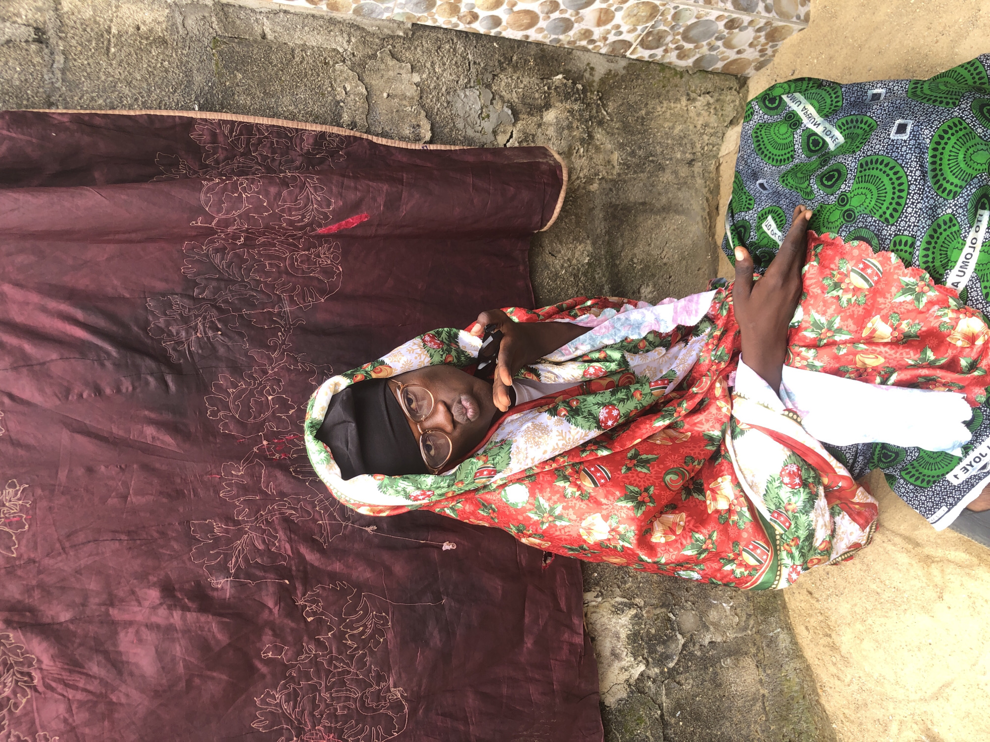 In Ado, Nigeria, a person sits on sandy ground in front of a textured, rust-colored fabric backdrop with embroidered floral motifs. The individual is wearing a black head covering and glasses, with a vibrant red, white, and green patterned garment draped over their head and shoulders. A white garment is visible underneath. They are also wearing a green and black patterned fabric wrapped around their lower body. The person is leaning forward, pursing their lips as if blowing a kiss. To the right of the backdrop, a section of a rough, grey concrete wall is visible, interspersed with decorative tiles featuring circular patterns. The overall scene suggests an outdoor setting within a residential area in Ado.