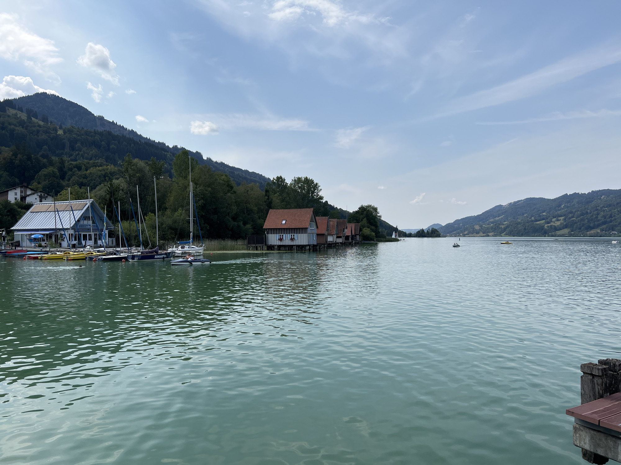 This image depicts a scenic view of the Alpsee lake, located in Immenstadt im Allgäu, Germany. The foreground is dominated by the tranquil, blue-green waters of the lake, with gentle ripples reflecting the sky. Along the left bank, several boats are moored, including sailboats and smaller recreational vessels, adjacent to a building with a distinctive blue awning and a white roof, likely a boat house or club facility. Further along the shore, a row of traditional wooden boathouses with red tiled roofs are visible. In the background, rolling green hills covered in dense forests rise towards a sky with scattered white clouds. The overall atmosphere is serene and idyllic, suggesting a pleasant day at the lake.