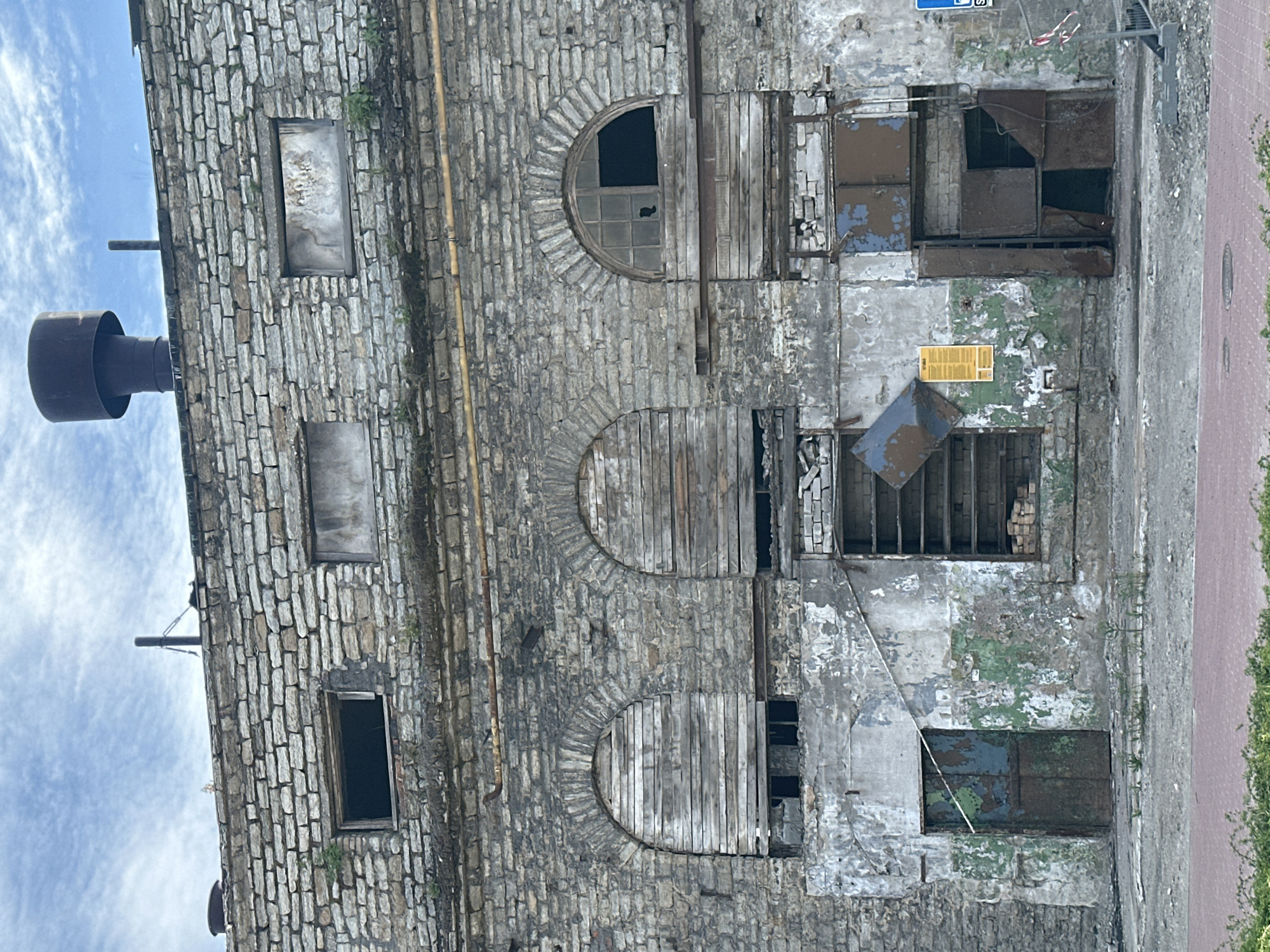 The image depicts the exterior of a derelict, multi-story building constructed of rough-hewn, grey stone blocks. The building appears to be located in Tallinn, Estonia, given the architectural style and the presence of similar industrial structures in the city. The facade is weathered and damaged, with peeling paint and vegetation growing from cracks in the stonework.
Several windows are visible. Some are boarded up with wooden planks, while others are broken or empty, revealing dark interiors. A large arched window on the right has a broken pane. Two arched openings higher up are also covered with wooden slats.
At the ground level, there are entrances that appear to be doors, but they are heavily rusted and some are partially open, showing dark openings. One door on the left is a single, rusted metal panel. To the right of this, there is a larger, more complex metal entryway with multiple panels, some of which are bent or detached, revealing the interior. A yellow sign is affixed to the wall above this area.
On the rooftop, a large, dark, cylindrical chimney or vent is visible, along with a smaller vertical pipe. The sky above is cloudy with patches of blue. The ground in front of the building is a mix of rough asphalt, gravel, and some sparse vegetation. Red paving stones are visible in the lower foreground, suggesting a sidewalk or pedestrian area. Construction barriers and tape are present on the right side of the image, indicating ongoing work or a restricted area. No people are visible in the image.