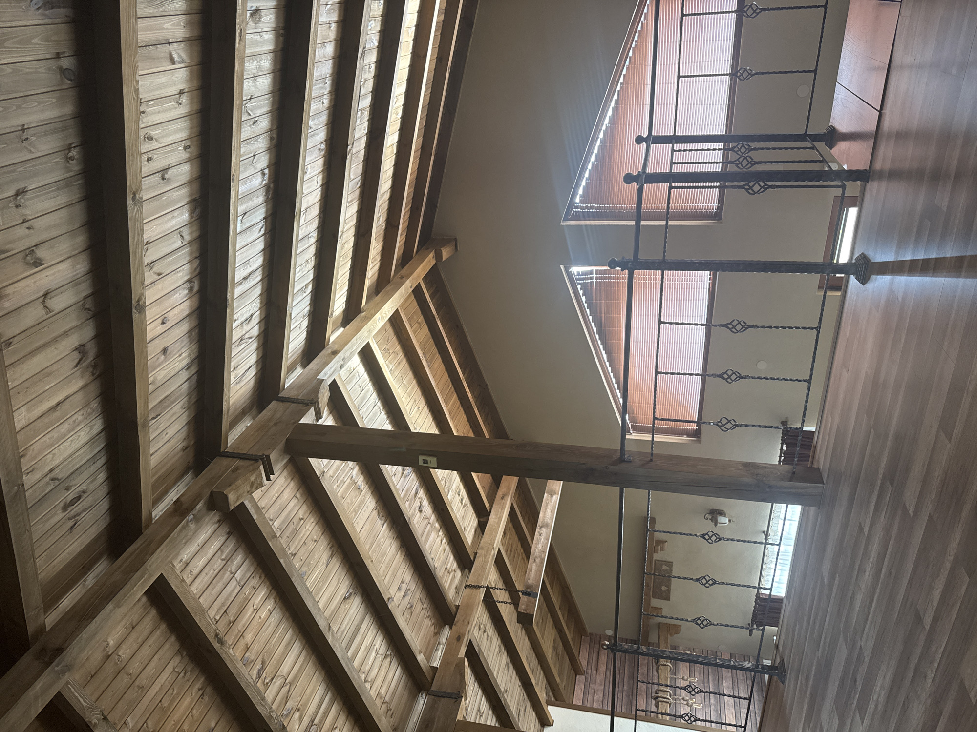 A photograph taken indoors, likely in Dolni Marian, Bulgaria, presents a distorted perspective looking upward toward the ceiling. The composition is bisected, with a steeply angled wooden structure dominating the left side. This structure comprises parallel wooden planks and supporting beams, exhibiting a rustic, unfinished aesthetic. The right side of the image reveals an interior space with a wooden floor and what appears to be a railing with ornate ironwork. Windows are present, covered by horizontal blinds, which diffuse the incoming light. The ceiling is a neutral color, providing a visual separation between the wooden structure and the interior space. The angle of the photograph creates a sense of disorientation.