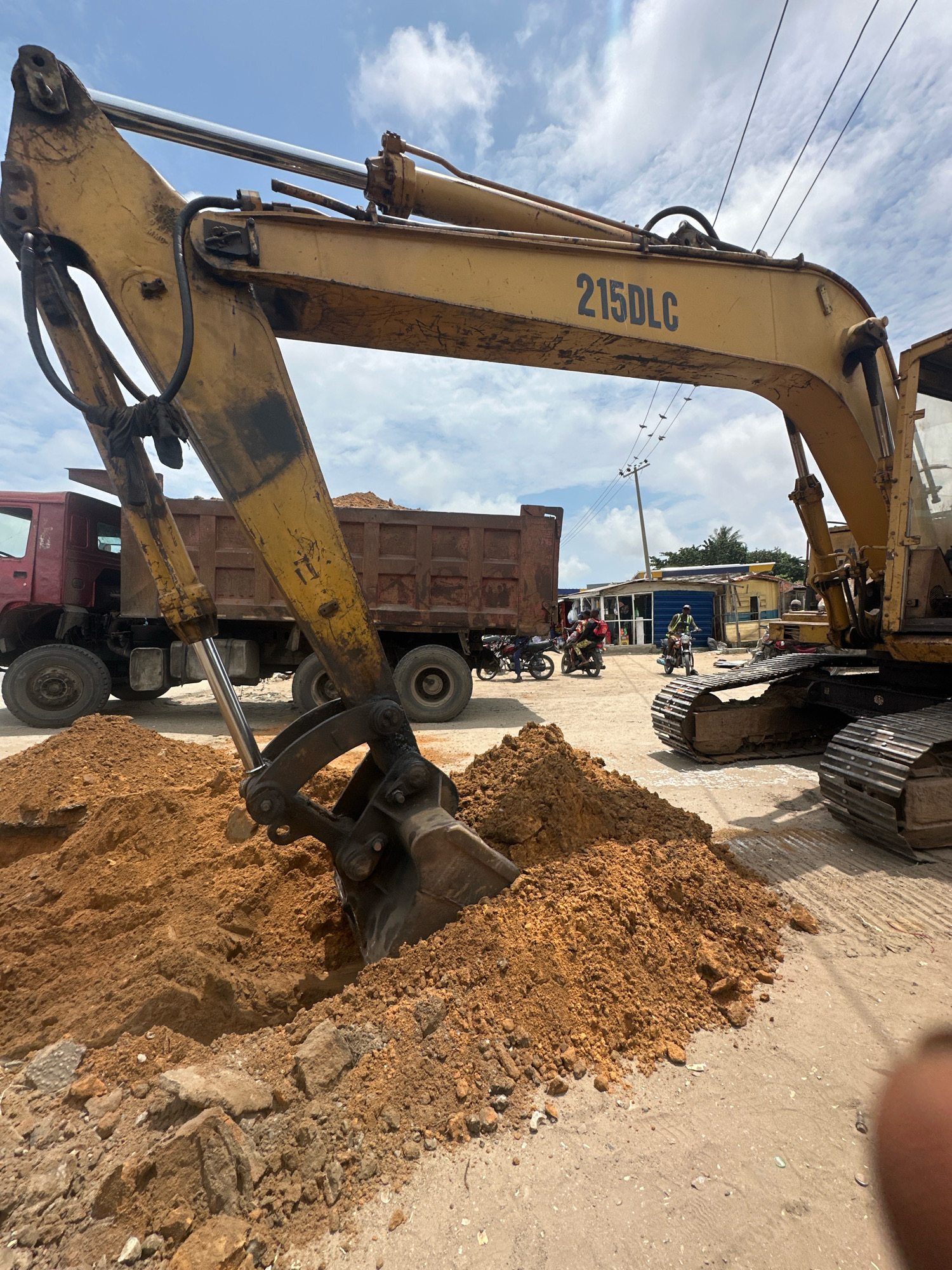In Ajara, Nigeria, an excavator is actively digging a trench. The excavator's bucket is partially submerged in the disturbed earth, with a pile of soil beside it. Behind the excavator, a large dump truck is parked, its bed also filled with soil. Several motorcycles are present, some with riders, suggesting ongoing activity in the area. In the background, buildings line a street, and overhead power lines are visible against a cloudy sky.