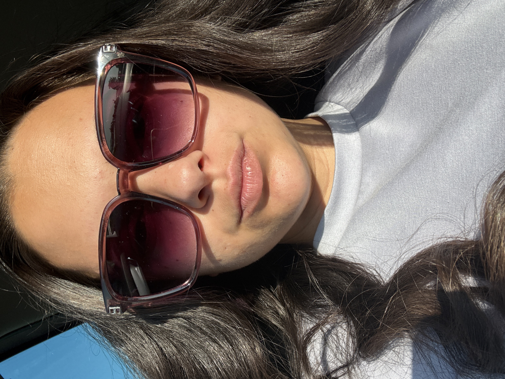 A woman is captured in a close-up, selfie-style shot, her face partially obscured by large, square, plum-colored sunglasses. Her dark, wavy hair frames her face, falling over her shoulders and onto her light blue t-shirt. The sunlight casts strong shadows on her skin and clothing, creating a dramatic effect. The background is blurred, with hints of a car's interior and a bright blue sky visible through a window on the left. There is no identifiable information to place this image within Sliven, Bulgaria.
