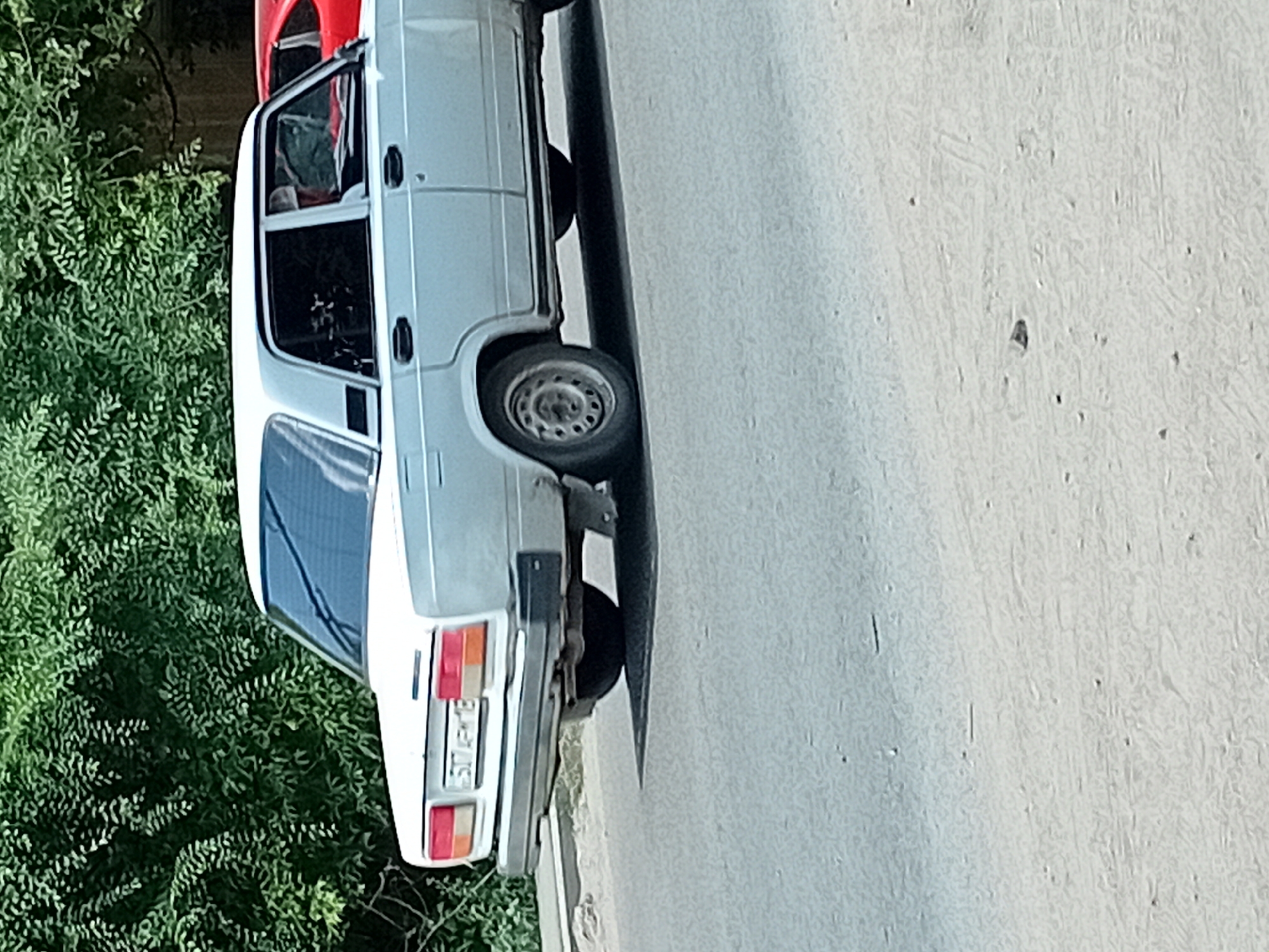 A light blue sedan is parked on an asphalt road, seen from its rear left side. The car has a white roof and black trim around the windows and doors. The license plate is visible and reads "517 APM 18". Behind the sedan, partially obscured by trees and vegetation, is a red car. The environment appears to be outdoors in a city setting, possibly Semey, Kazakhstan, indicated by the license plate format. The road surface is grey asphalt with some debris. The lighting suggests it is daytime.