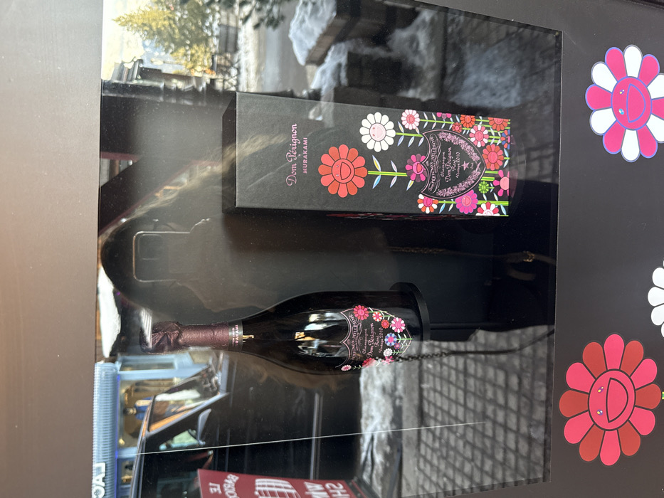 The image depicts a Dom Pérignon x Takashi Murakami champagne display within a retail or hospitality setting in Courchevel, France. A bottle of "Dom Pérignon Rosé Vintage 2010" with a colorful floral Murakami label and a corresponding black gift box are presented inside a transparent display case. The case surface reflects a person taking the photograph, visible as a silhouette holding a smartphone, with long dark hair. The reflections also show a dark vehicle, a building with blue accents (possibly a shop with partial text "LE BAROQ" visible), and an outdoor winter environment featuring snow-covered ground, wooden structures, and evergreen trees, confirming the snowy mountain resort context of Courchevel. Two Murakami flower stickers, one red and one pink/white, are adhered to the lower front of the display case.