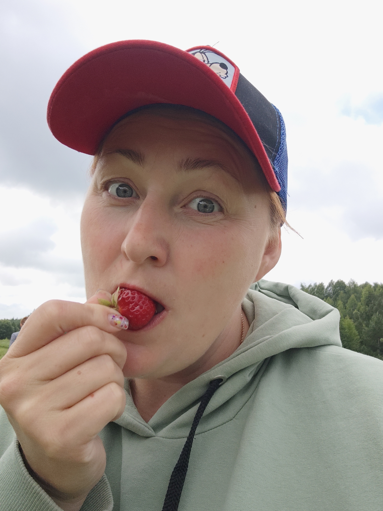 A woman is seen from the chest up, outdoors in what appears to be a rural setting with trees in the background and a cloudy sky. She is wearing a light green hooded sweatshirt and a red and blue baseball cap. She is holding a strawberry up to her mouth, about to eat it, and looking directly at the camera with a surprised or curious expression. Her fingernails are decorated with a colorful, patterned design. The image is a close-up, with the background slightly blurred. The specific location in Zhigalki, Russia, cannot be determined from the visual information provided.
