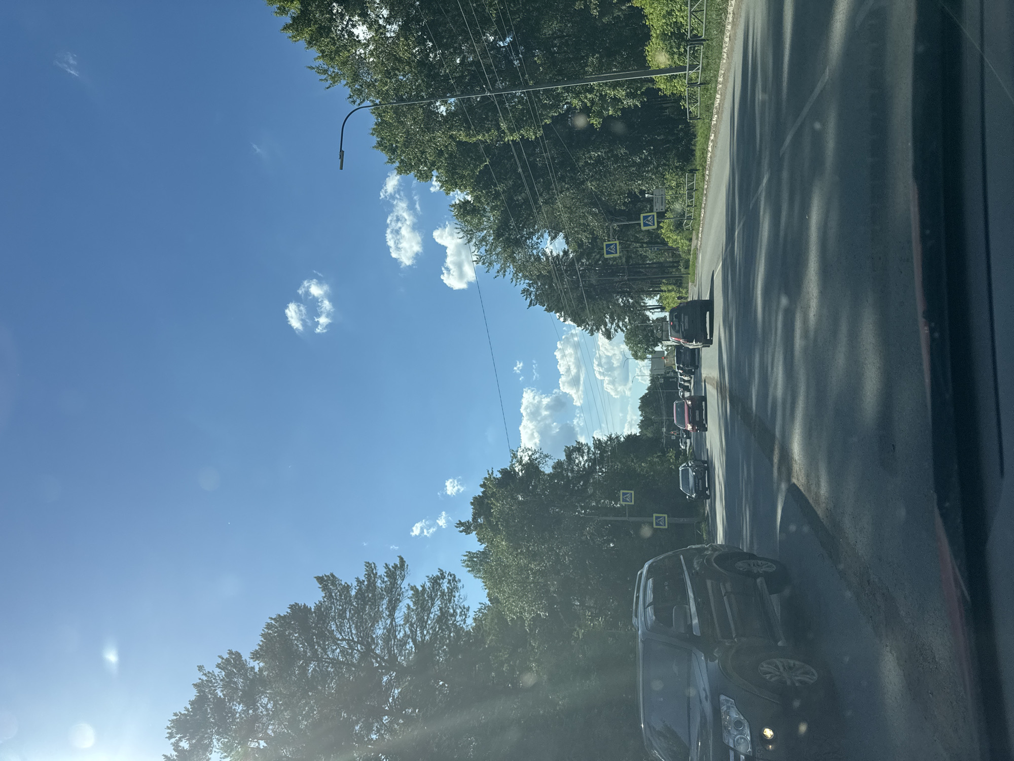 The image depicts a sunny day on a two-lane road lined with dense trees on both sides. The sky is bright blue with scattered white clouds. Several cars are visible on the road, moving in the same direction. A black SUV is in the foreground on the left, and a red car is further ahead on the right. Pedestrian crossing signs are posted along the road. The scene appears to be a suburban or semi-rural area, possibly on the outskirts of Chaikovsky, Russia, given the presence of trees and what looks like a traffic light in the distance. The overall impression is of normal daytime traffic under clear weather conditions.