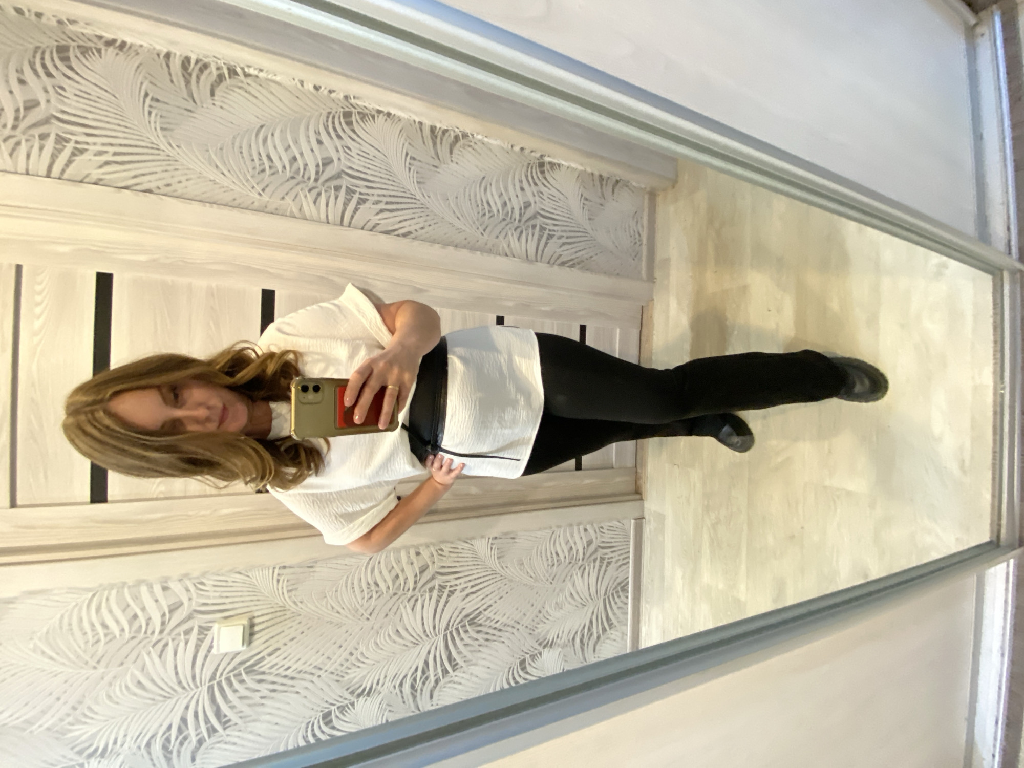 A woman is taking a selfie in front of a mirror, wearing a white shirt and black pants. She is holding a smartphone in her right hand, and her left hand is on her hip. Her blonde hair is shoulder-length. The mirror reflects a portion of a room with patterned wallpaper and a light-colored floor. The city and location are not verifiable from the image.