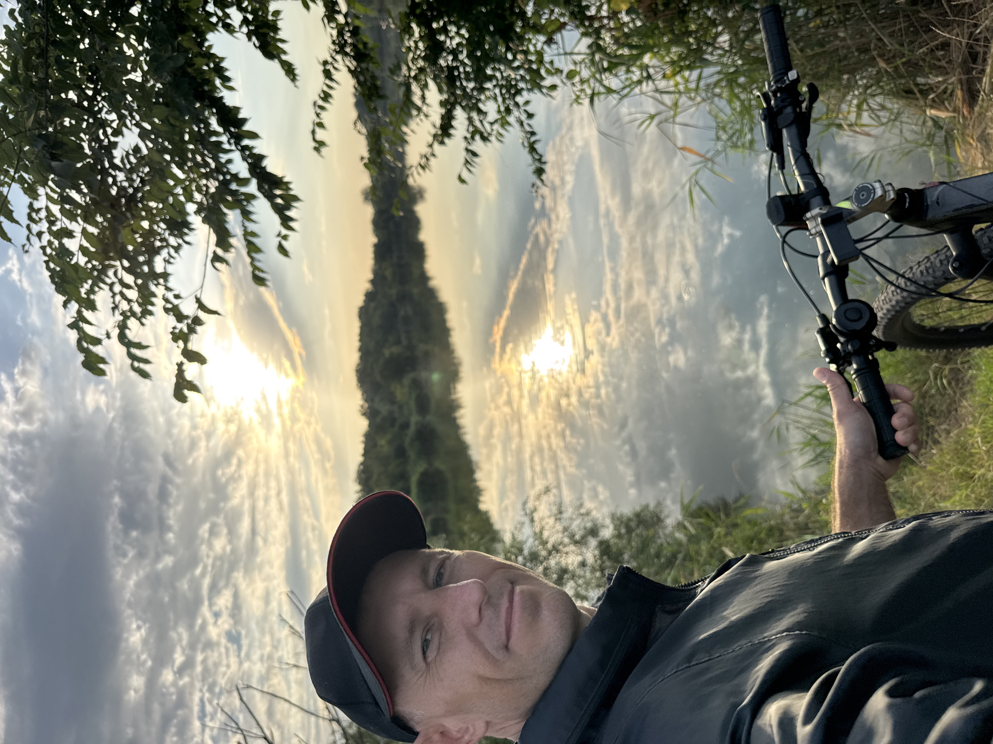 A man wearing a black baseball cap and a black jacket is taking a selfie outdoors by a body of water. He is positioned on the left side of the frame, looking towards the camera with a slight smile. The sun is setting or rising, casting a warm glow on the clouds and reflecting on the calm water. Trees and reeds line the background and foreground, with the handlebars and front wheel of a bicycle visible in the lower right quadrant of the image, suggesting he is on a bike ride. The environment appears to be natural and tranquil.
