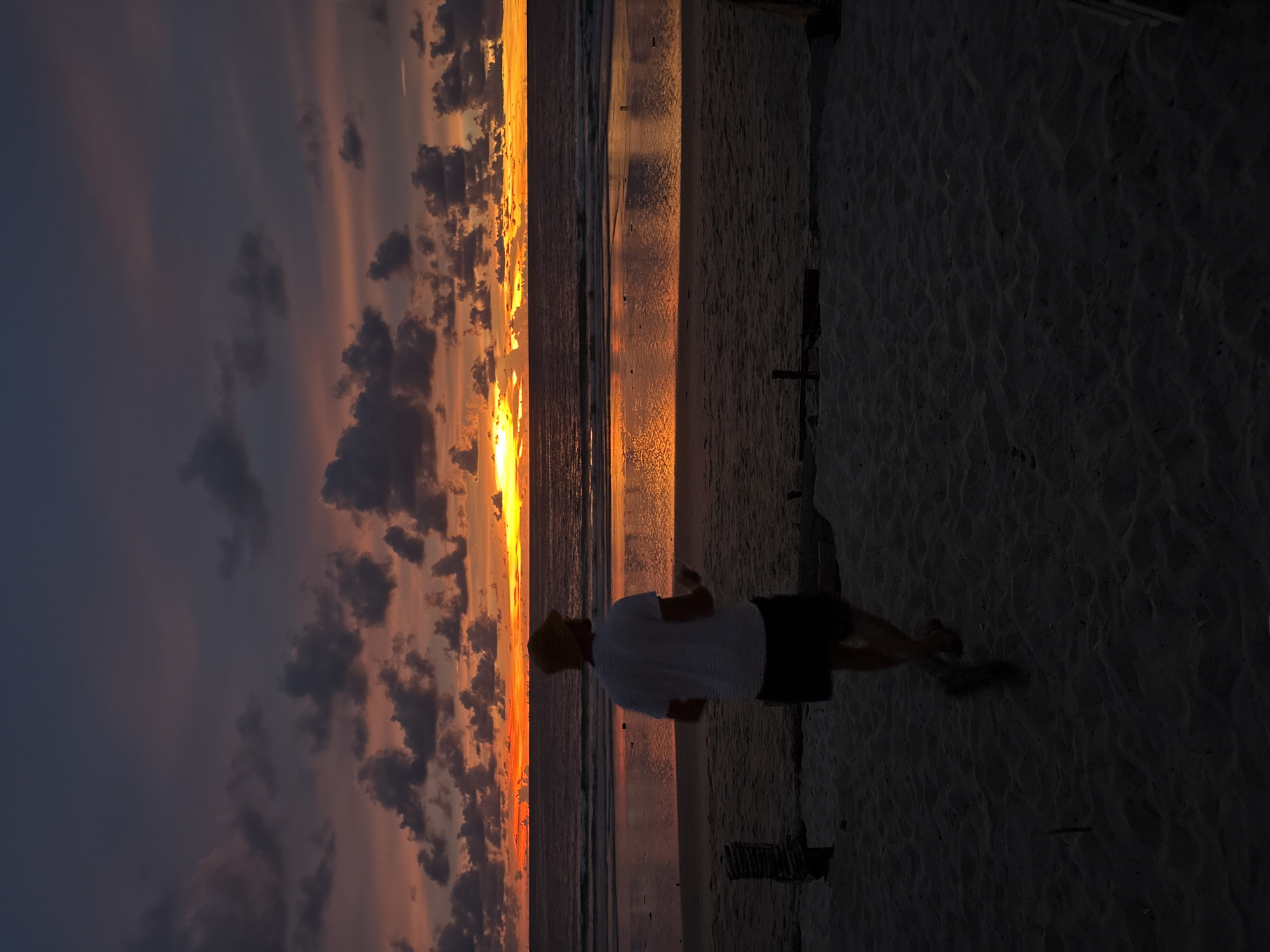 A person, silhouetted against a vibrant sunset, is walking along a sandy beach. The sky is a dramatic display of orange, yellow, and purple hues, with dark clouds adding texture. The ocean reflects the sunset's colors, creating a shimmering pathway. The sand is a deep brown, with ripples and footprints suggesting recent activity.
The person is wearing a white t-shirt and dark shorts, with a yellow hat. Their silhouette is the primary focus in the foreground of the image. The overall mood is peaceful and serene, with the natural beauty of the sunset dominating the scene. The time of day is clearly sunset. The location appears to be a tropical beach, possibly in Southeast Asia, given the warm tones and the presence of palm trees in the distance (though not clearly visible in this crop). There is no visible text in the image.