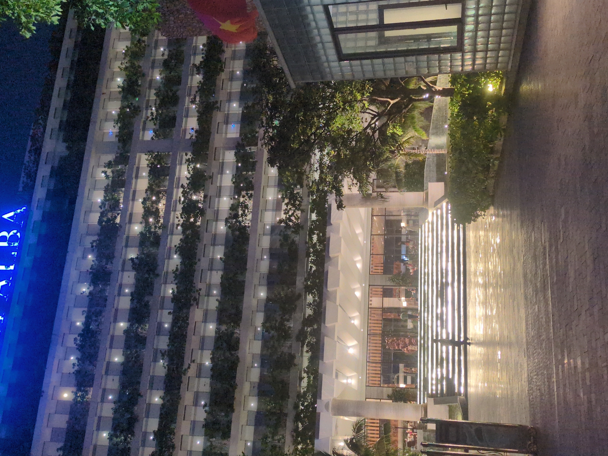 Nighttime view of a modern building in Tuy Hòa, Vietnam. The building's facade is decorated with vertical green strips and illuminated windows. A large, blue sign reading "ALBA" is partially visible on the left side of the building. Trees and plants surround the building, and a brick pathway leads up to the entrance. A red Vietnamese flag is placed near the top of the building. The area is well-lit, and the reflection of lights can be seen on the pathway.