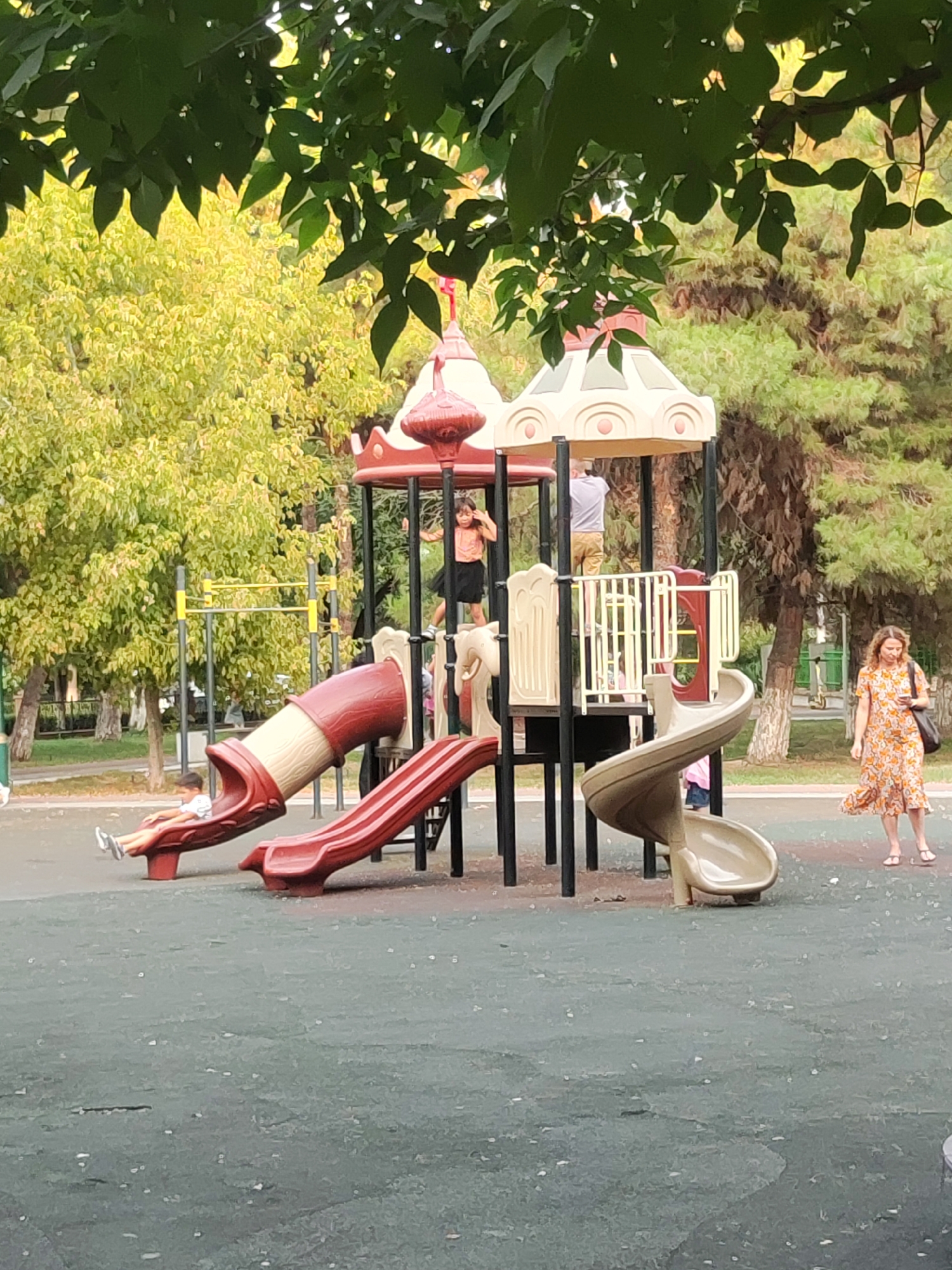 This image depicts a playground scene in Tashkent, Uzbekistan, under a canopy of green leaves. The focal point is a multi-level play structure with red and beige plastic elements, including slides and platforms. On the left side of the play structure, a young boy in a white t-shirt and shorts is positioned on a red slide, appearing to be resting or preparing to slide. Further up on the play structure, a young girl in a peach-colored top and black skirt stands on a platform. Another individual, a man in a light gray t-shirt and tan shorts, is also on the play structure, seemingly looking at something above. To the right of the play structure, a woman with curly brown hair, wearing a patterned orange dress and white sandals, walks away from the viewer, carrying a dark bag over her shoulder and looking at her phone. The playground is situated on a dark green, rubberized surface. In the background, there are numerous trees with lush green foliage and some pine trees. Yellow playground equipment, possibly monkey bars, is visible behind the main play structure. The overall environment suggests a park or recreational area within a city setting.