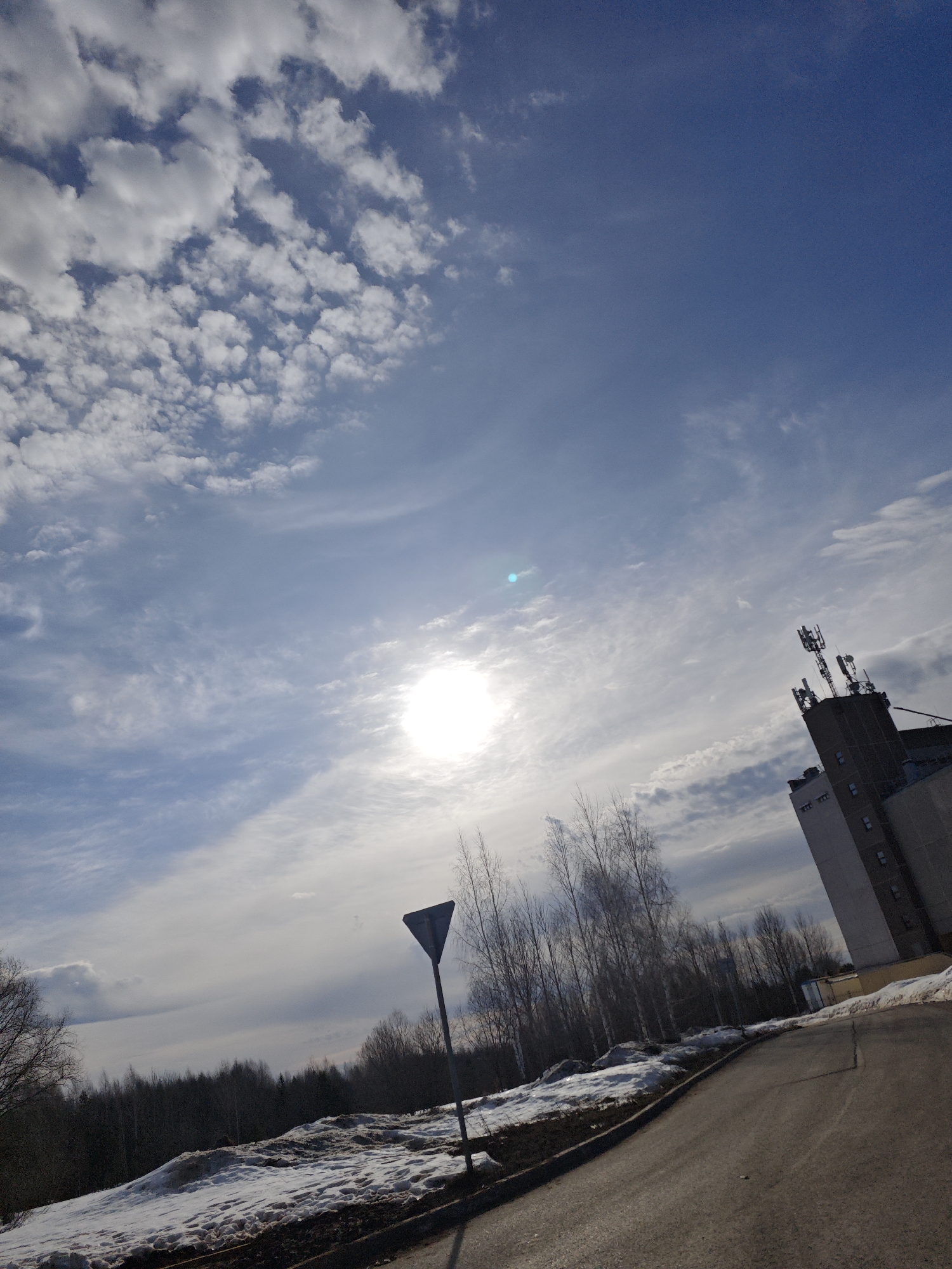 The image depicts an outdoor scene in the city of Марковский, Russia, on a bright day with a partially cloudy sky. The sun is prominently visible in the upper center, producing significant glare and a greenish-blue lens flare. The sky above is a clear blue, transitioning to a lighter hue near the horizon, populated by scattered cumulus clouds on the left and wispy cirrus formations stretching across the frame.
In the foreground, an asphalt road curves from the lower right towards the middle left. Adjacent to the left edge of the road, patches of melting snow, mixed with dirt, cover the ground. A dark, triangular traffic sign, likely a Yield sign, is mounted on a pole near the road on the left. Behind the snow patches and the sign, a line of bare deciduous trees, possibly birches, stands on an elevated bank, followed by a dense forest extending into the background.
On the right side of the image, a tall, multi-story building, appearing to be constructed of light gray concrete, rises into the sky. Its flat roof is equipped with multiple communication antennas and dishes. A section of the building's base is painted yellow. No individuals or active interactions are visible within the frame. The overall impression is of a late winter or early spring environment.