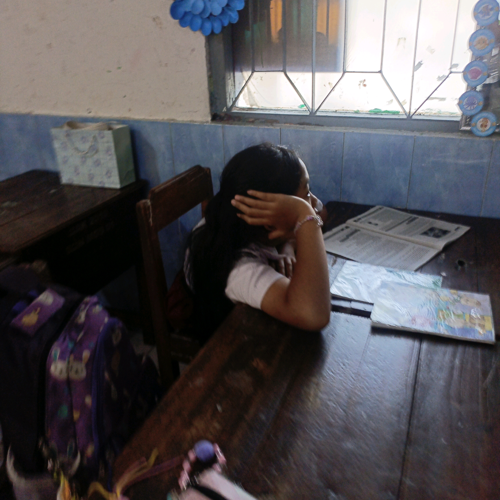 A young girl with dark hair is sitting at a wooden desk, her head resting on her hand, and she is looking to the right. She is wearing a white t-shirt. There are two books on the desk in front of her. To her left, there is a purple backpack with a cat pattern. In the background, there is a window with blue tiled walls and a light-colored wall above. A blue paper decoration is hanging from the top left corner of the image. There are also some decorations on the window grid. The scene appears to be in a classroom in Salatiga, Indonesia.