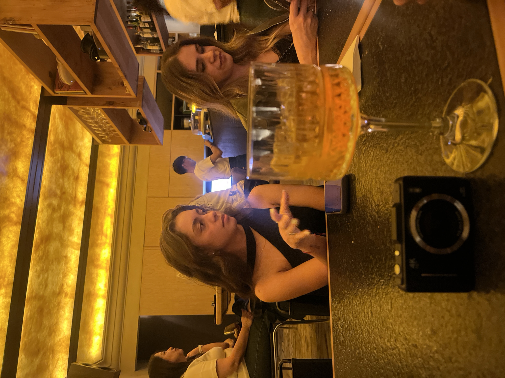 The image depicts two women sitting at a bar, with a glass of amber-colored liquid and a black camera prominently displayed on the counter. The woman closest to the camera is gesturing with her right hand. Another person is visible in the background. The location appears to be a bar with a modern aesthetic, featuring illuminated panels. There is no information given that confirms this image was taken in the city of San Bernardino Tlaxcalancingo, Mexico.