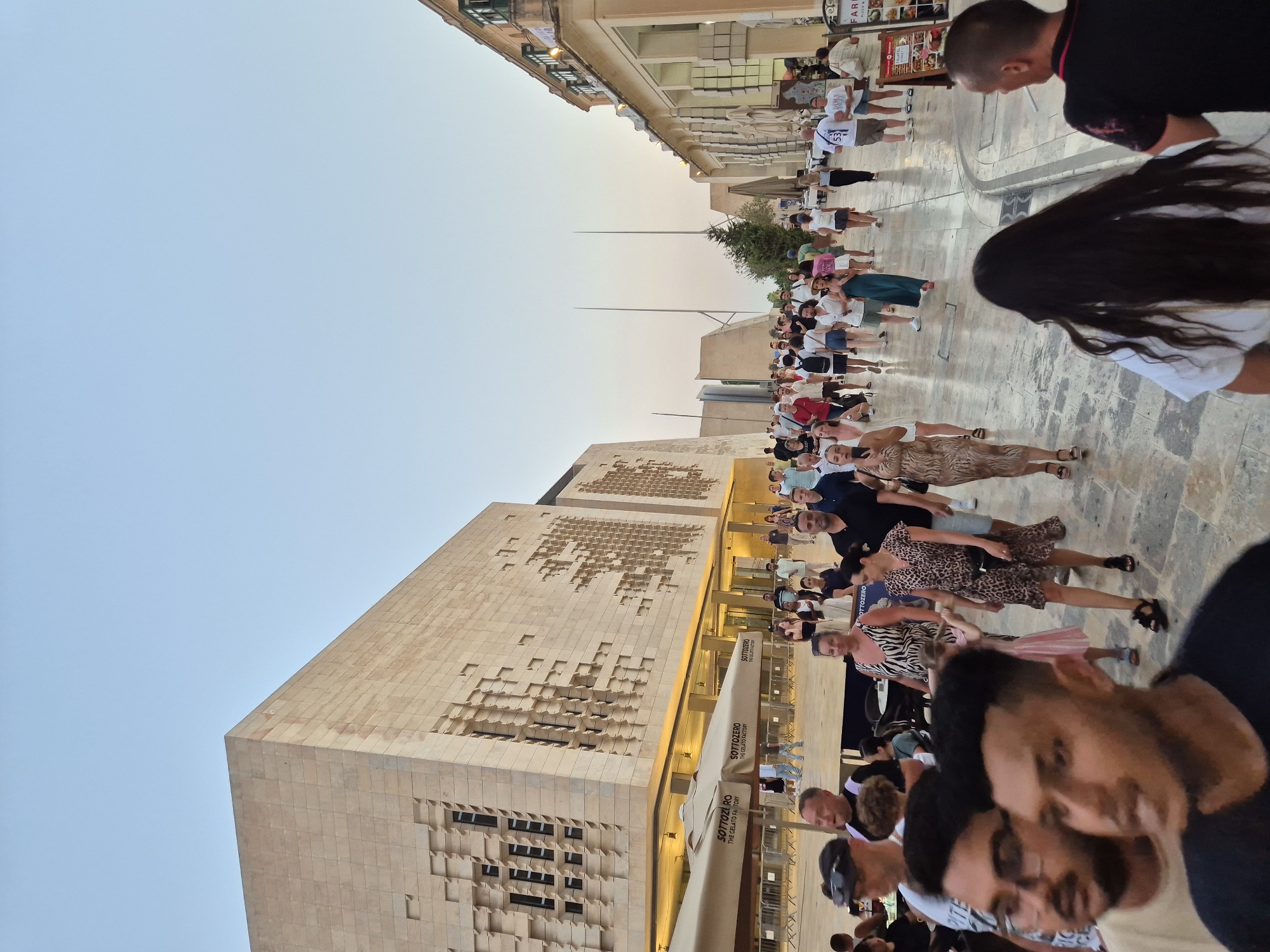 The photograph depicts a bustling street scene in Valletta, Malta, specifically in front of the Parliament House, a modern building with a distinctive geometric facade. The building's exterior is constructed from light-colored stone, with a series of recessed window-like openings creating a textured pattern. In the foreground, a diverse group of people are walking along a paved street. Their clothing suggests a warm climate, with many individuals wearing casual summer attire. Several people are looking towards the camera, while others are engaged in conversation or walking in various directions. Large outdoor umbrellas, branded with "SOTTOZERO THE GELATO FACTORY," are visible to the left of the frame, indicating the presence of a food establishment. The street is lined with buildings on the right, featuring traditional Maltese architecture with balconies. The sky is clear and light blue, suggesting either early morning or late afternoon. The overall impression is one of a lively urban environment during a period of good weather.